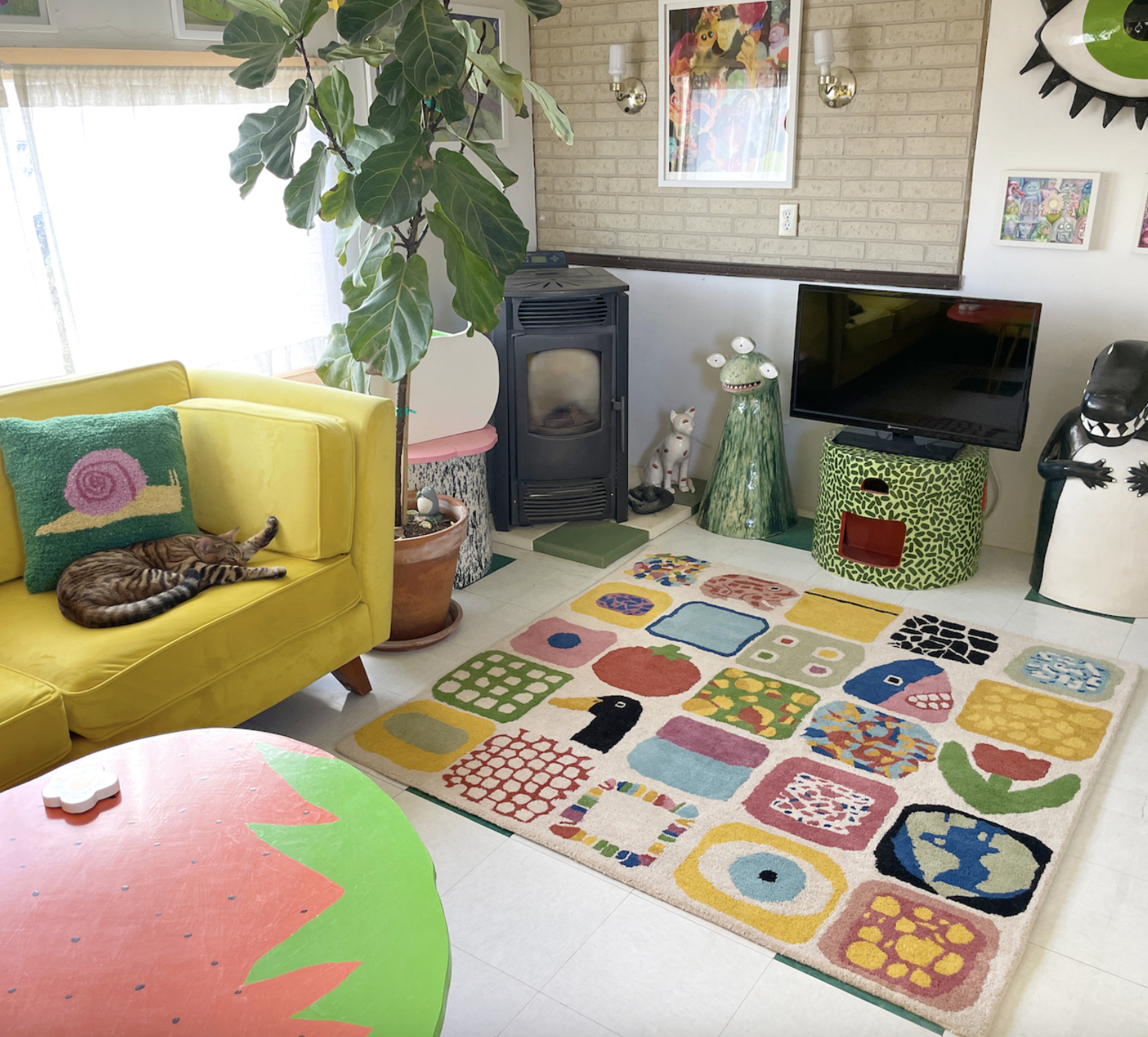 Living room with yellow sofa and a tabby cat sleeping on it, colorful patterned rug, large houseplant, black stove, television on a green patterned stand, wall art and sculptures including a frog figure and a black and white figure, and a pink and green table in the foreground.