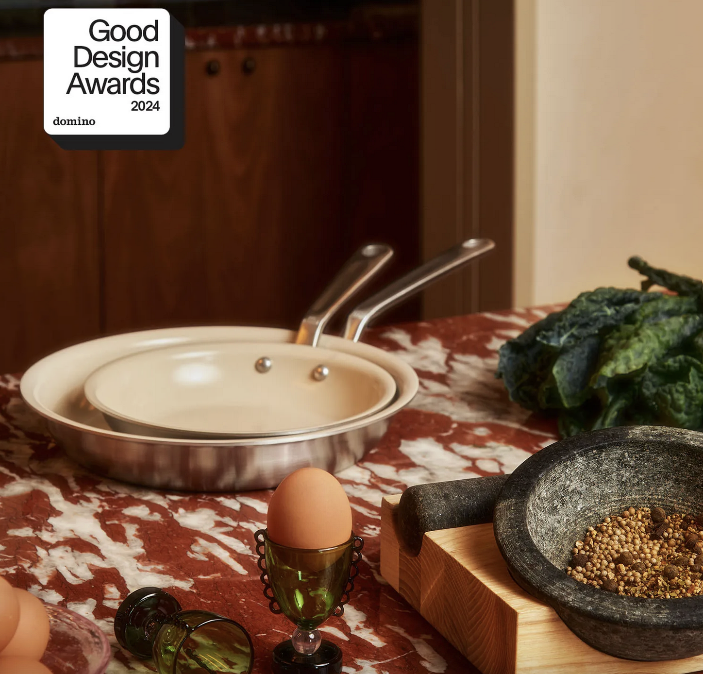 Kitchen counters with cookware, including a large frying pan, a smaller pan, a stone mortar and pestle with peppercorns, eggs in an egg cup, and greens, set on a red and white marble surface.