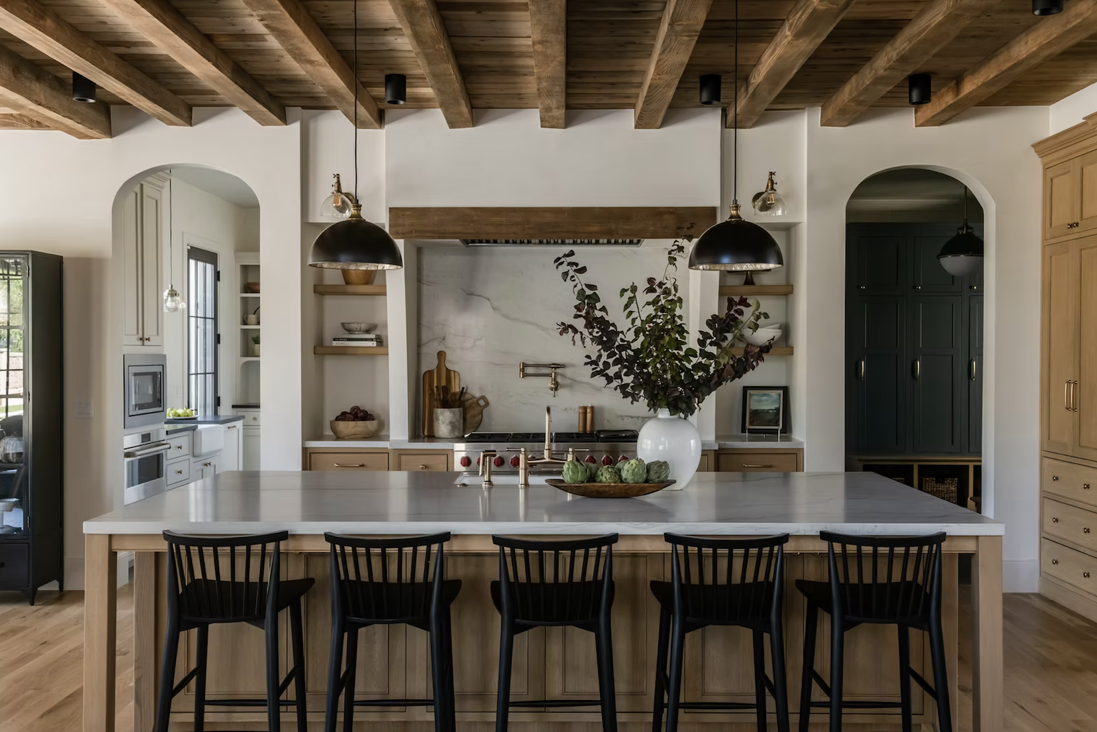 Modern kitchen with a central island, black chairs, wooden ceiling beams, and built-in shelves. Decor includes a large white vase with greenery and art pieces.