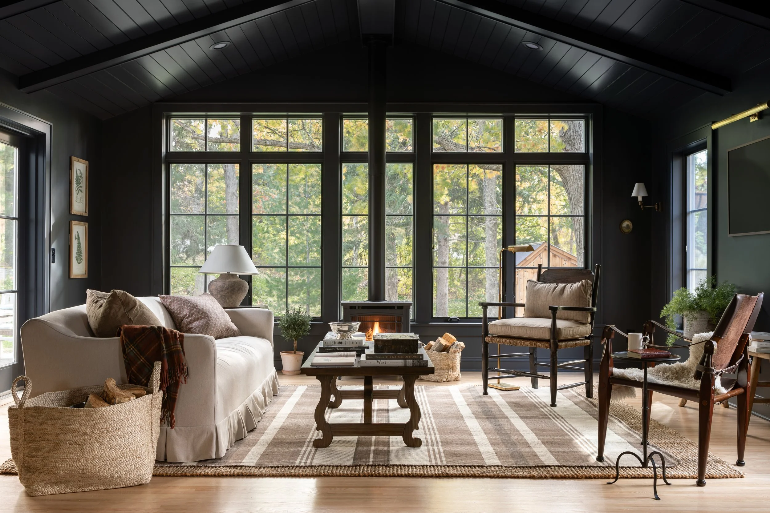 A cozy living room with large windows overlooking trees, furnished with a white sofa, various chairs, a wooden coffee table, a rug, and decorative items.