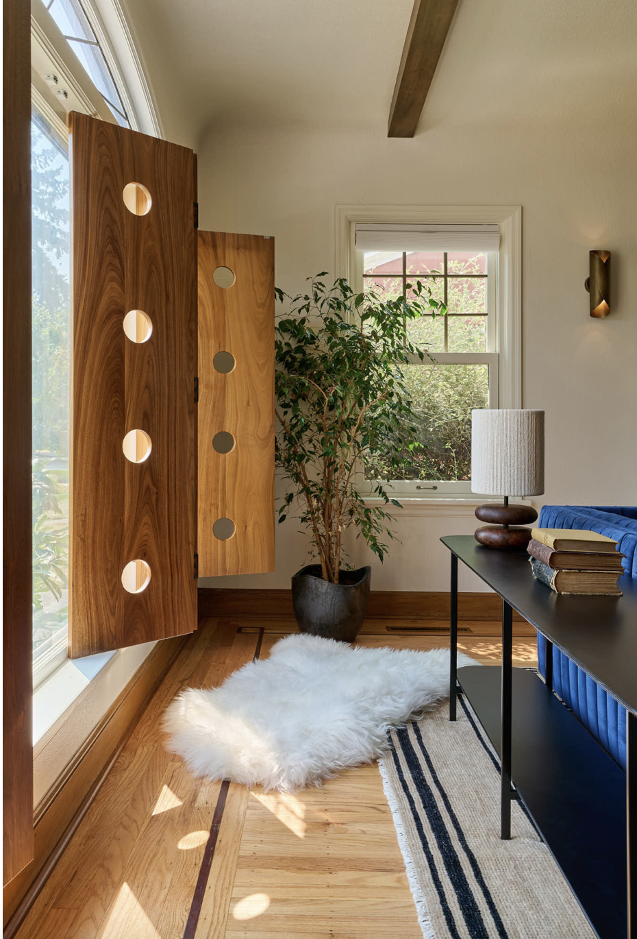 A cozy corner with a large window, a wooden privacy screen with round cutouts, a potted plant, a table lamp, stacked books, a soft white fur rug, and a striped rug on a wooden floor.