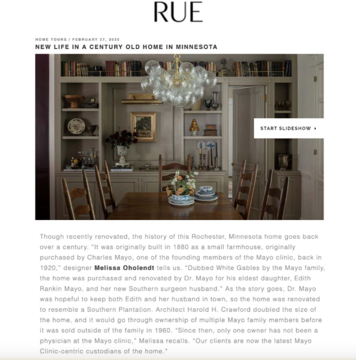 Interior of a dining room with a wooden table, six wooden chairs, a built-in bookshelf with books and decorative items, and a chandelier with glass globes hanging from the ceiling.