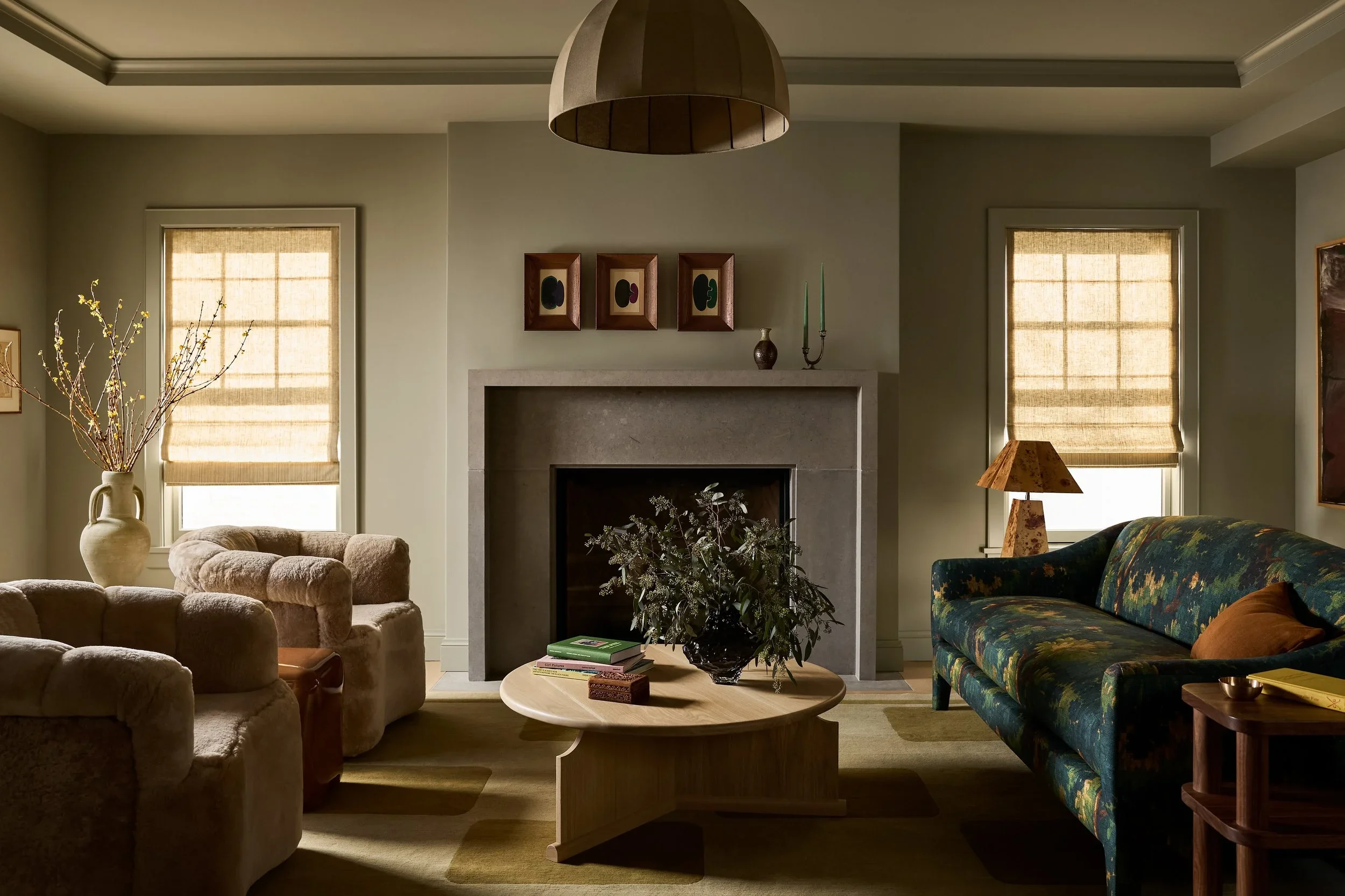 Living room with beige armchairs, a floral sofa, a round wooden coffee table with books and a plant, two windows with beige shades, and a fireplace with decorations above.