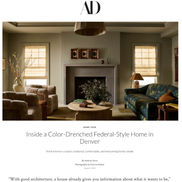 Interior of a color-drenched, Federal-style home in Denver showing a living room with a fireplace, beige and dark green armchairs, a wooden coffee table, window blinds, and a fireplace decorated with small art pieces and a plant.