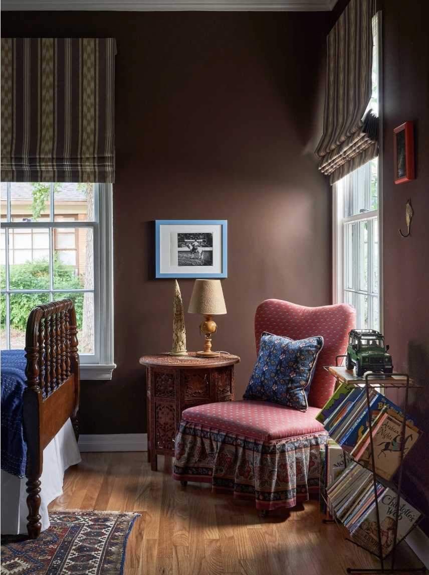 A cozy corner of a bedroom with a wooden bed, a side table, a table lamp, an upholstered chair with a pillow, a window with curtains, and a magazine rack.