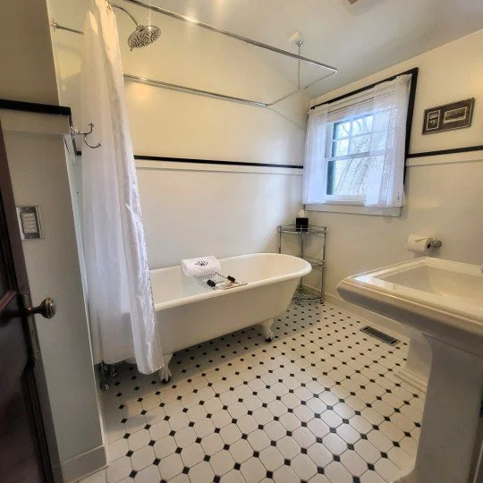 Bathroom with a white clawfoot bathtub, shower curtain, window with curtains, sink, small metal shelf, and black and white hexagonal tile flooring.