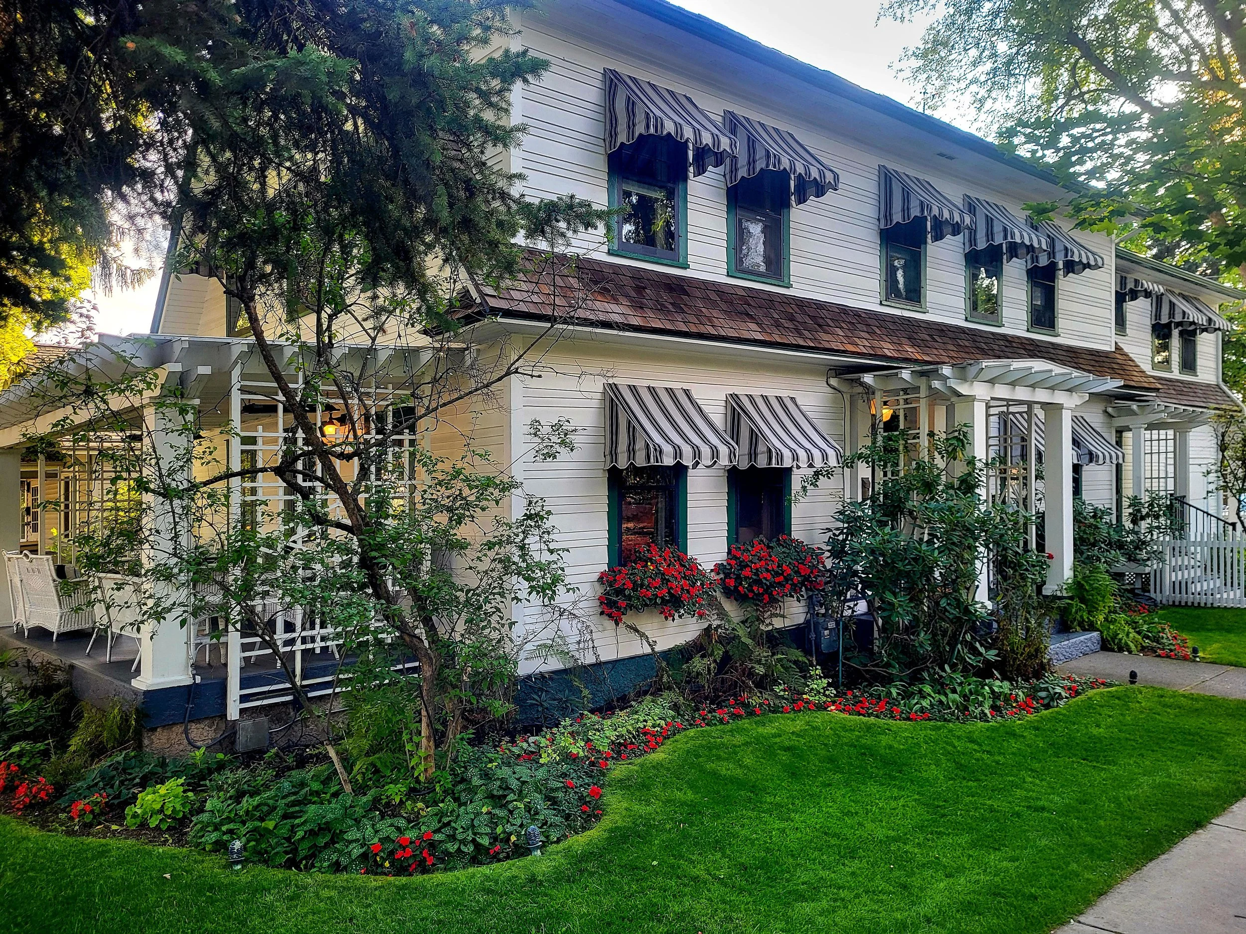 White two-story house with striped window awnings, a small porch with white pillars and a garden with red flowers, green grass, and trees around the house.