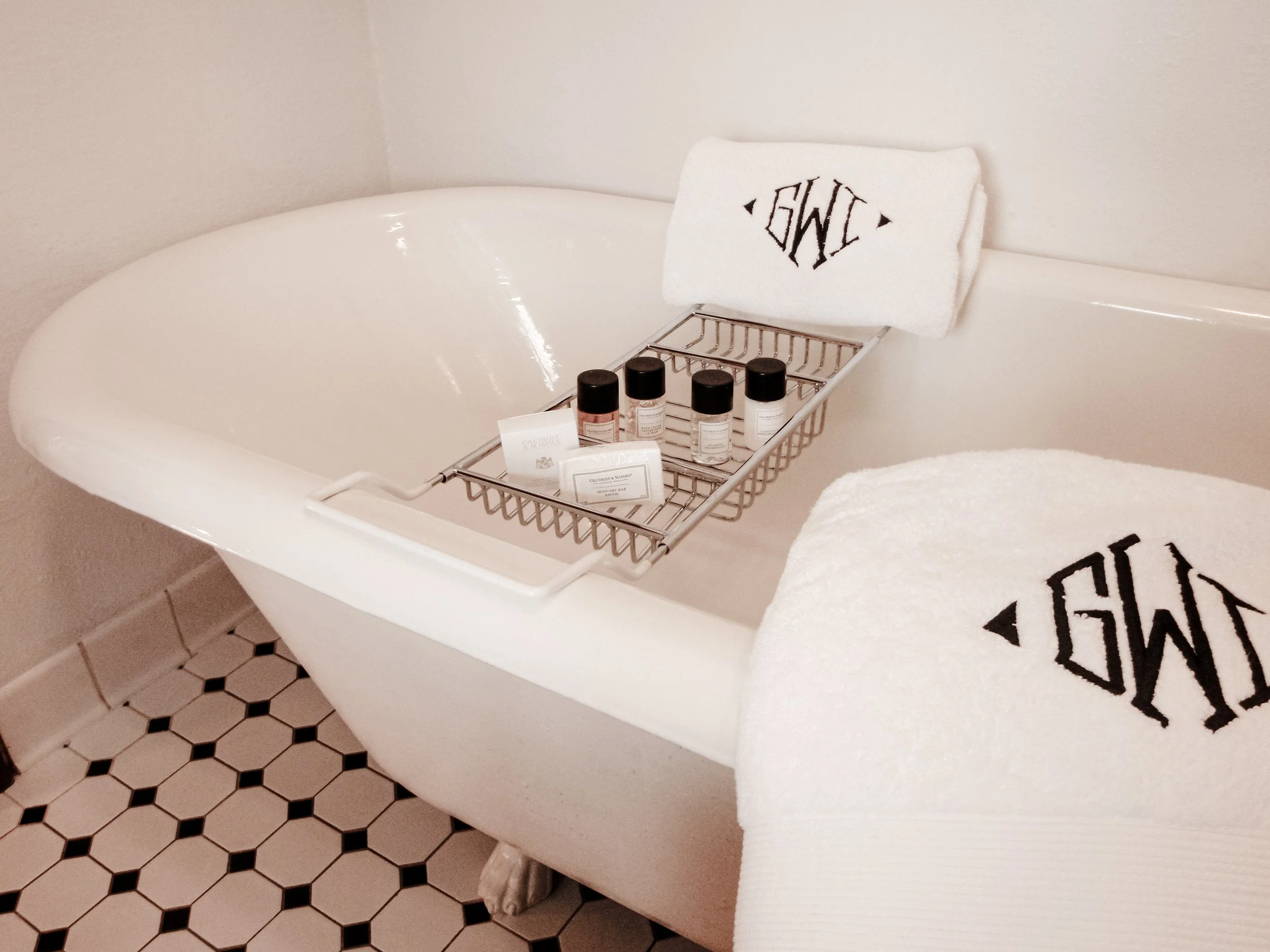 A white bathtub with a metal rack that holds four small bottles and packaged soap. There is a white towel with black embroidered letters folded on the edge of the tub. The floor has a black and white hexagonal tile pattern.