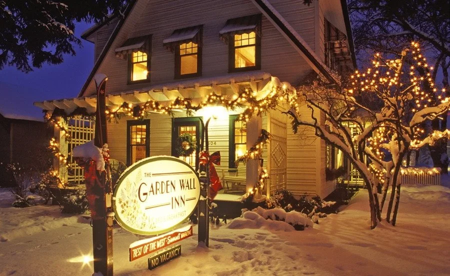 A warmly lit, decorated house during winter with snow on the ground and on trees, featuring holiday lights and a sign for The Garden Wall Inn.