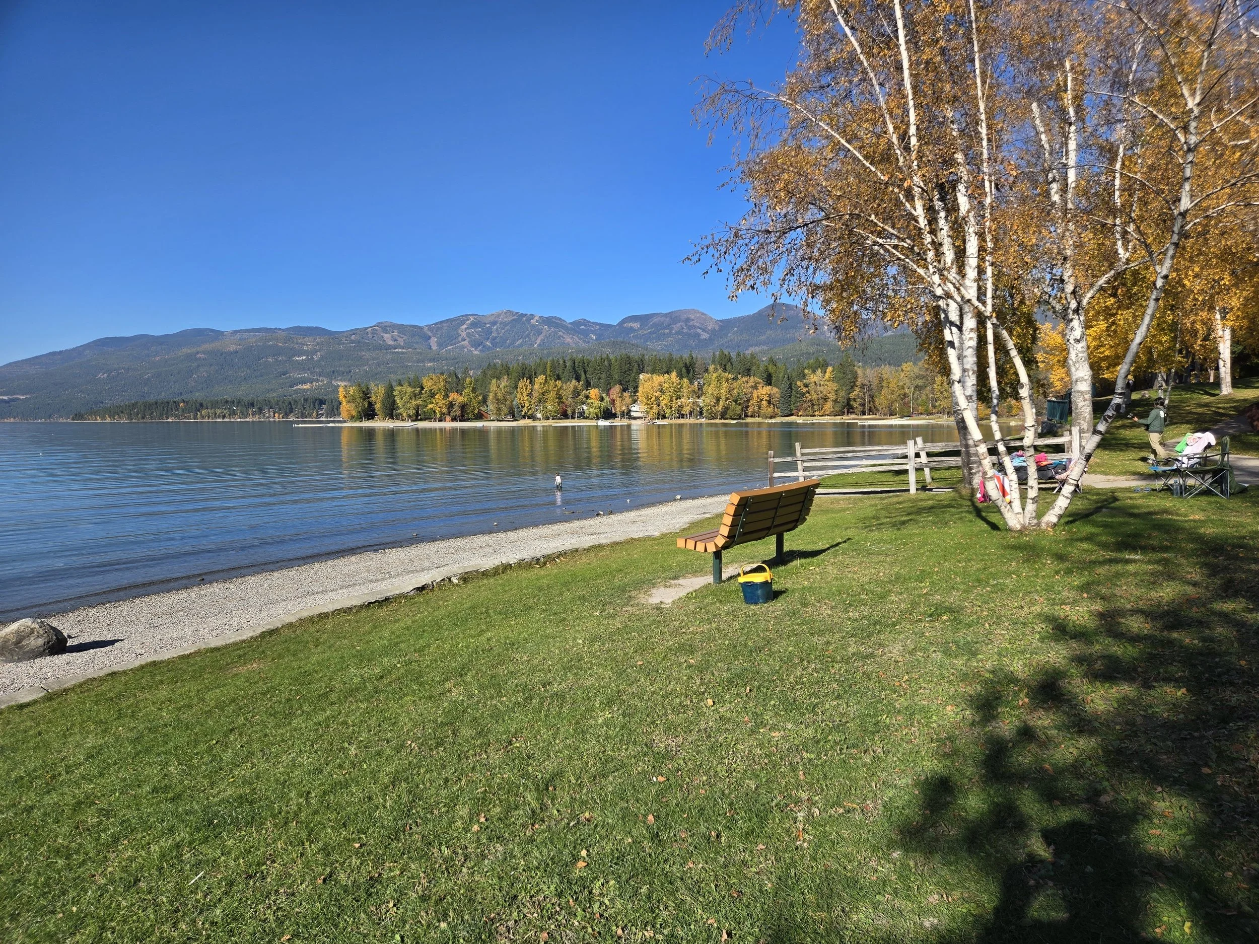 A peaceful lakeside scene at Whitefish Lake with mountain range in the background, and colorful autumn trees along the shore. 