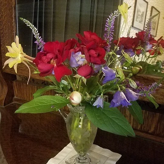 A floral arrangement in a tall clear glass vase with red, purple, yellow, and white flowers and green leaves, placed on a white cloth on a wooden surface.
