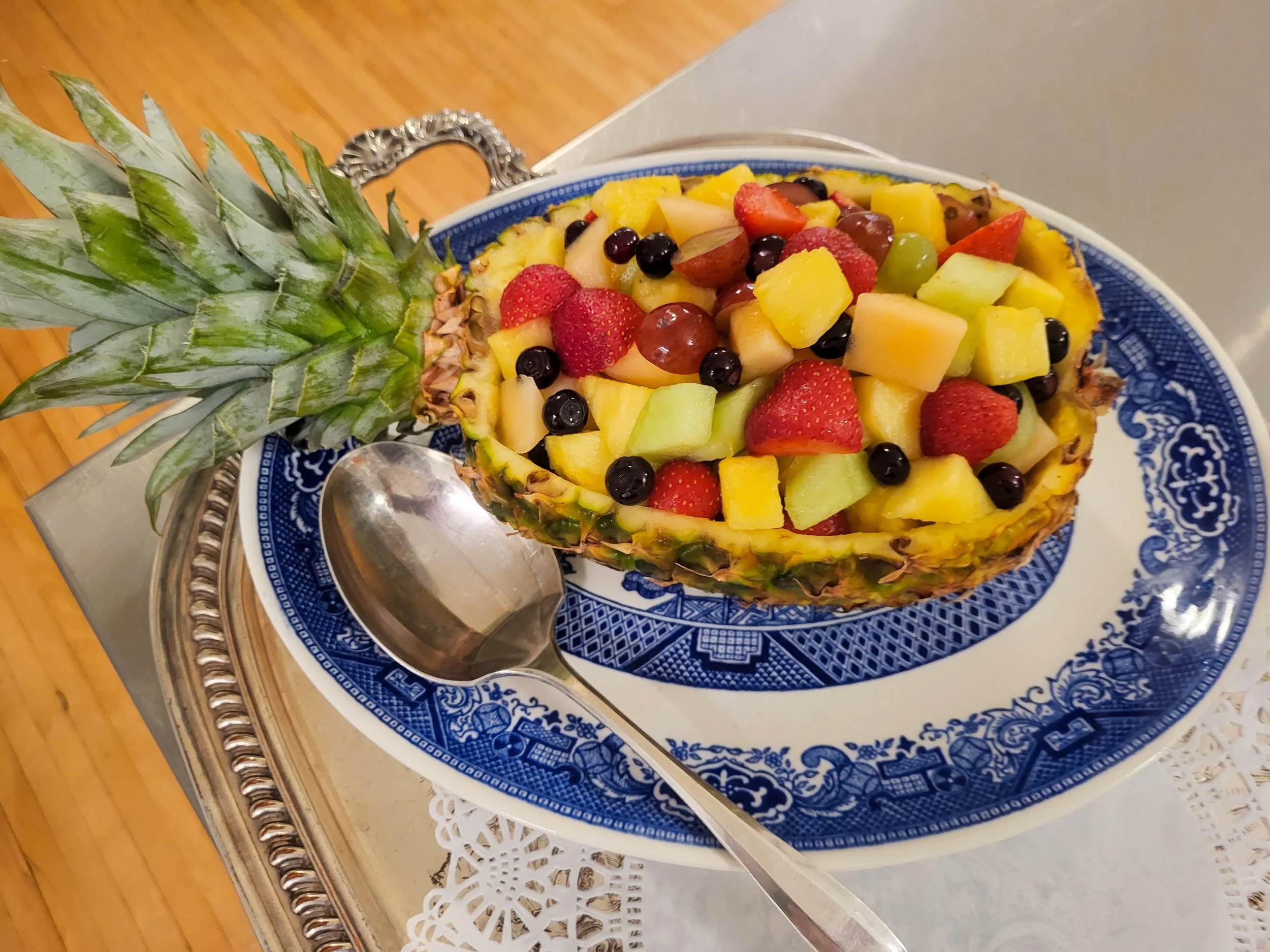 A halved pineapple filled with assorted fresh fruits including strawberries, blueberries, grapes, honeydew, cantaloupe, and pineapple chunks on a decorative blue and white plate.