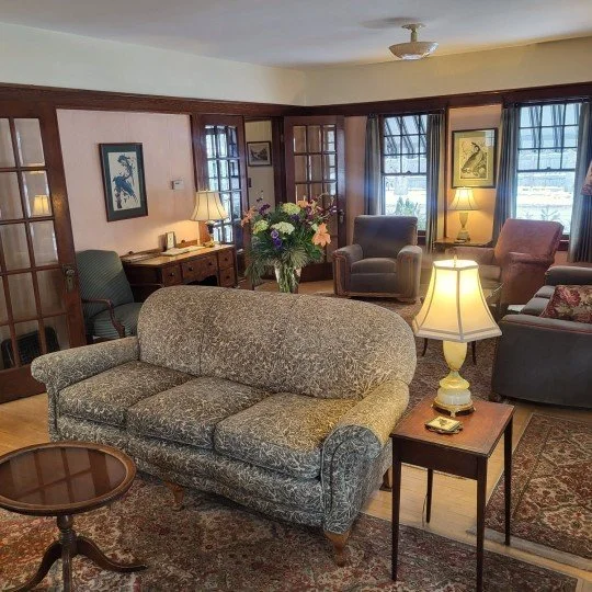 Living room with floral couch, side table with lamp, armchairs, wooden desk with lamp, and paintings on the walls.