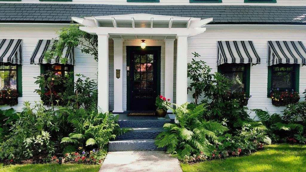 Front view of a white house with black door, striped awnings on windows, surrounded by plants and flowers.