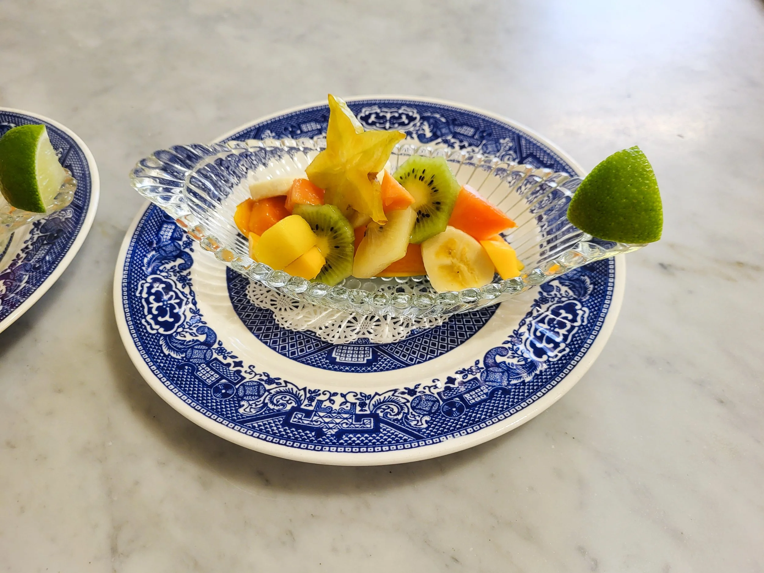 A small glass bowl with a decorative blue pattern containing chopped mixed fruit, including kiwi, pineapple, banana, and melon, on a blue and white patterned plate on a white marble surface.