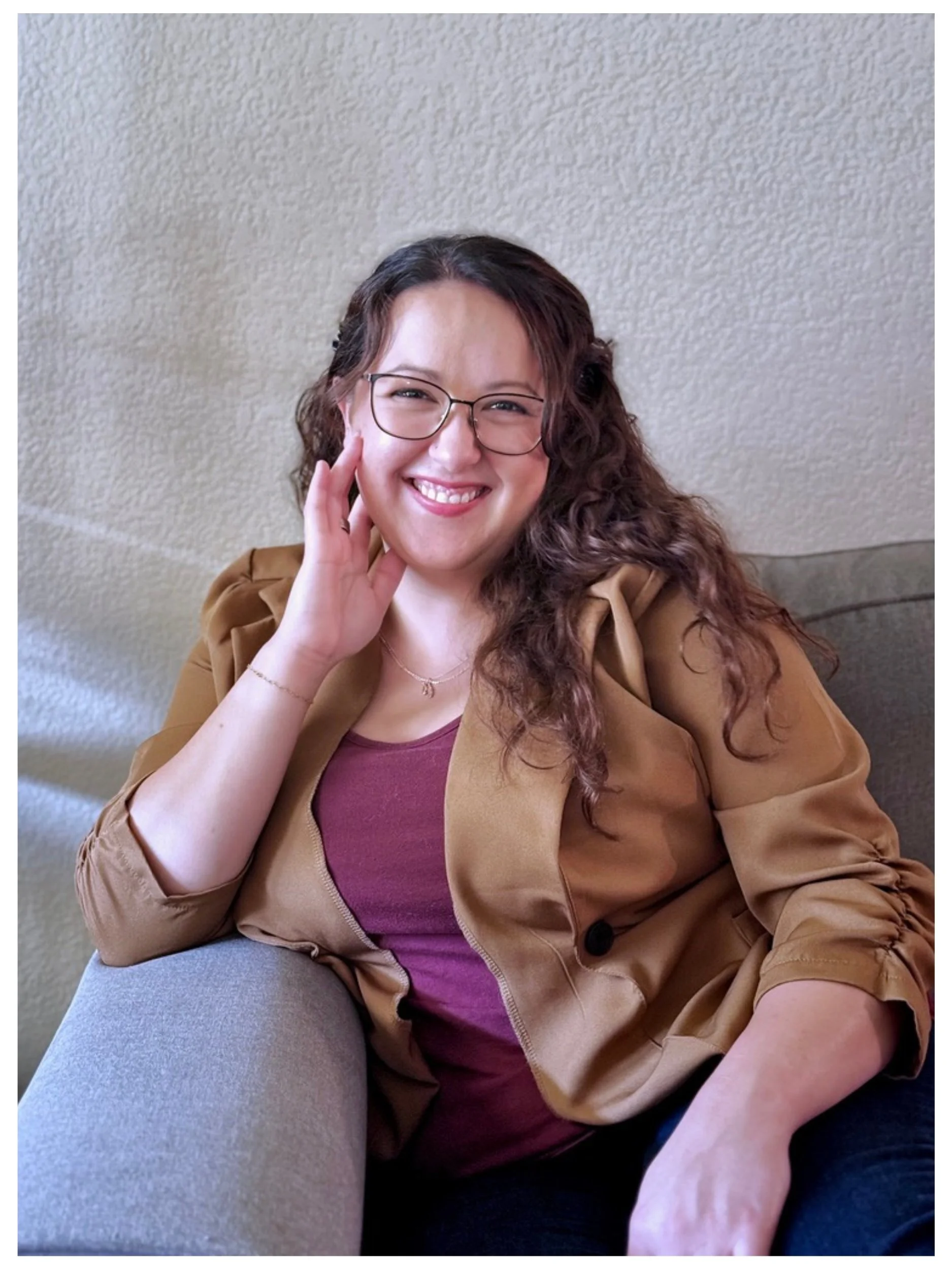 Smiling woman with glasses, long curly hair, wearing a tan blazer and maroon top, sitting on a gray couch against a textured beige wall.