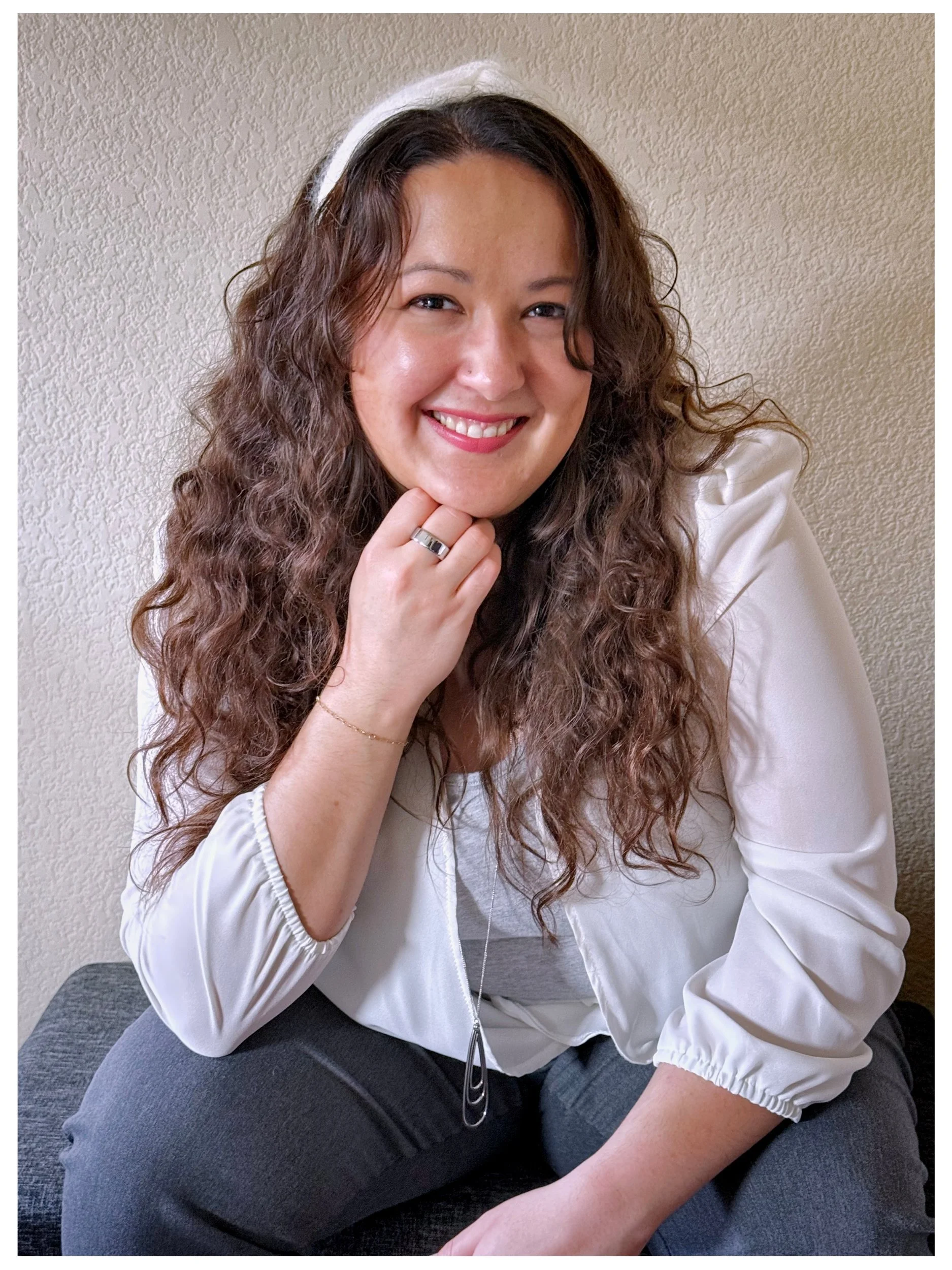 A woman with curly brown hair, wearing a white headband, white jacket, and gray pants, sitting on a gray upholstered surface, smiling and resting her chin on her hand.