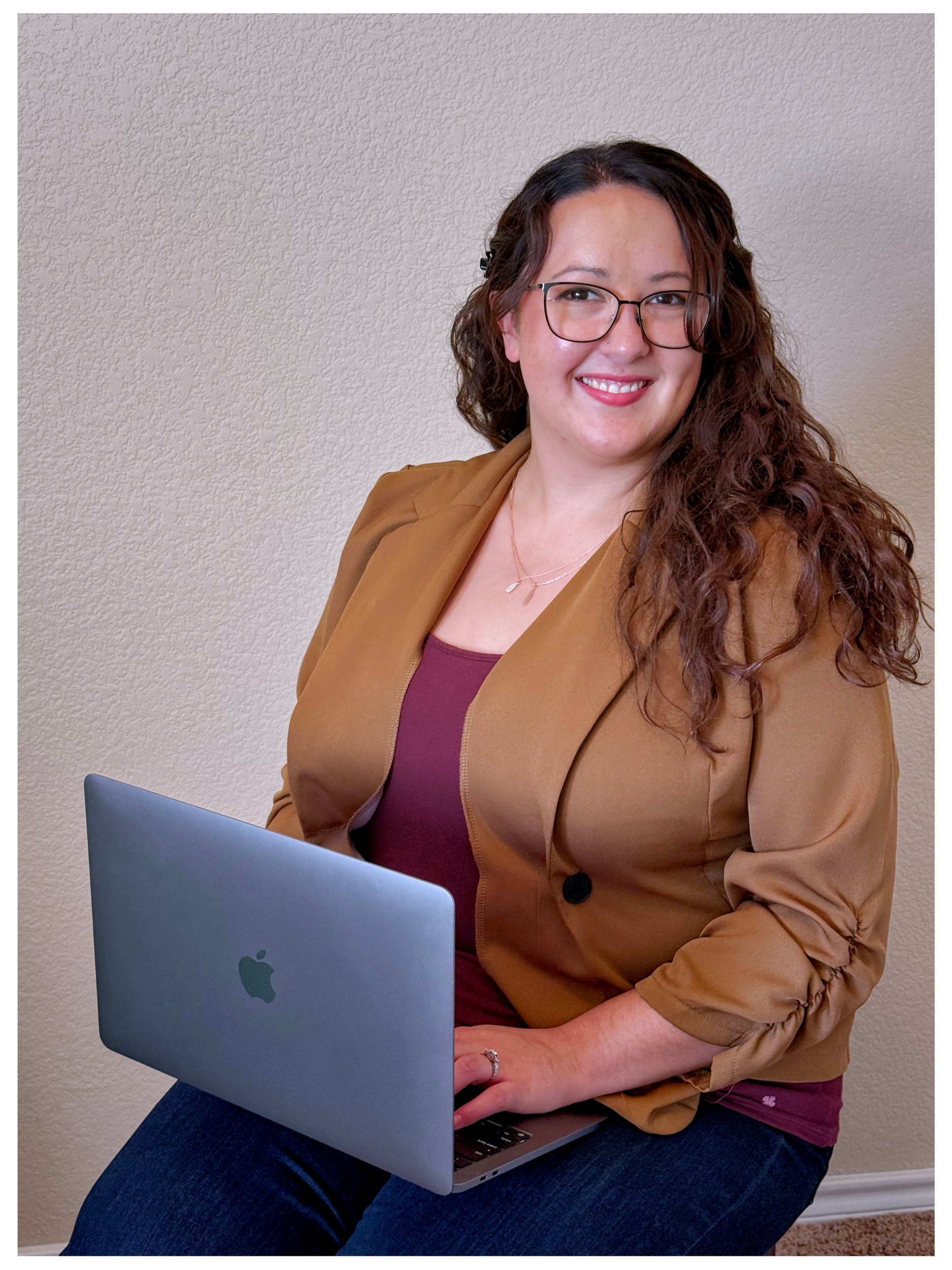 A woman with long curly brown hair, wearing glasses, a tan-colored blazer, and a maroon top, sitting and holding a silver MacBook laptop.