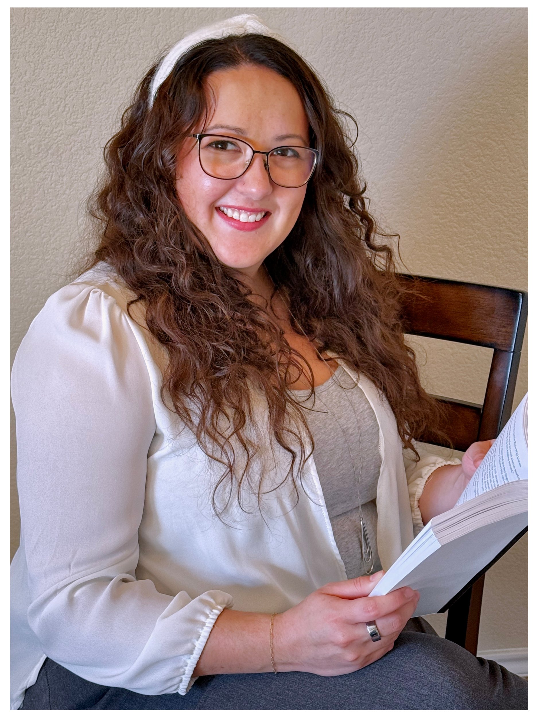 A woman with long, curly brown hair and glasses smiling while sitting and reading a book.