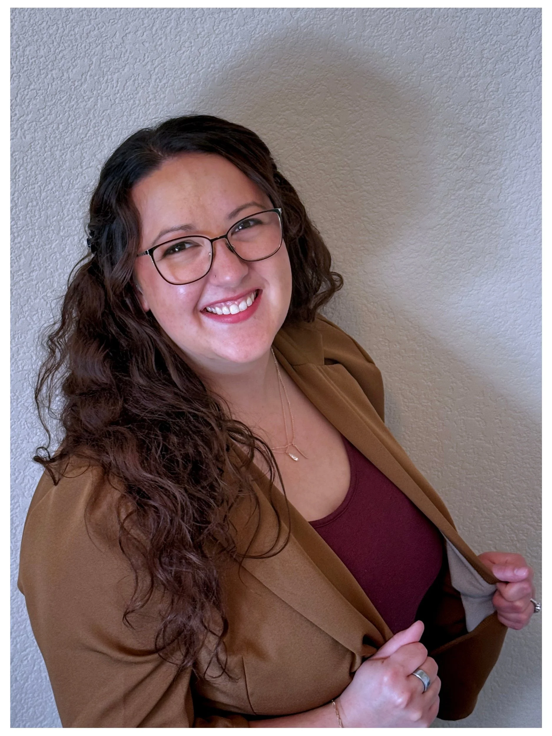A woman with long curly brown hair, glasses, and a bright smile, wearing a brown blazer over a maroon top, standing against a textured beige wall.