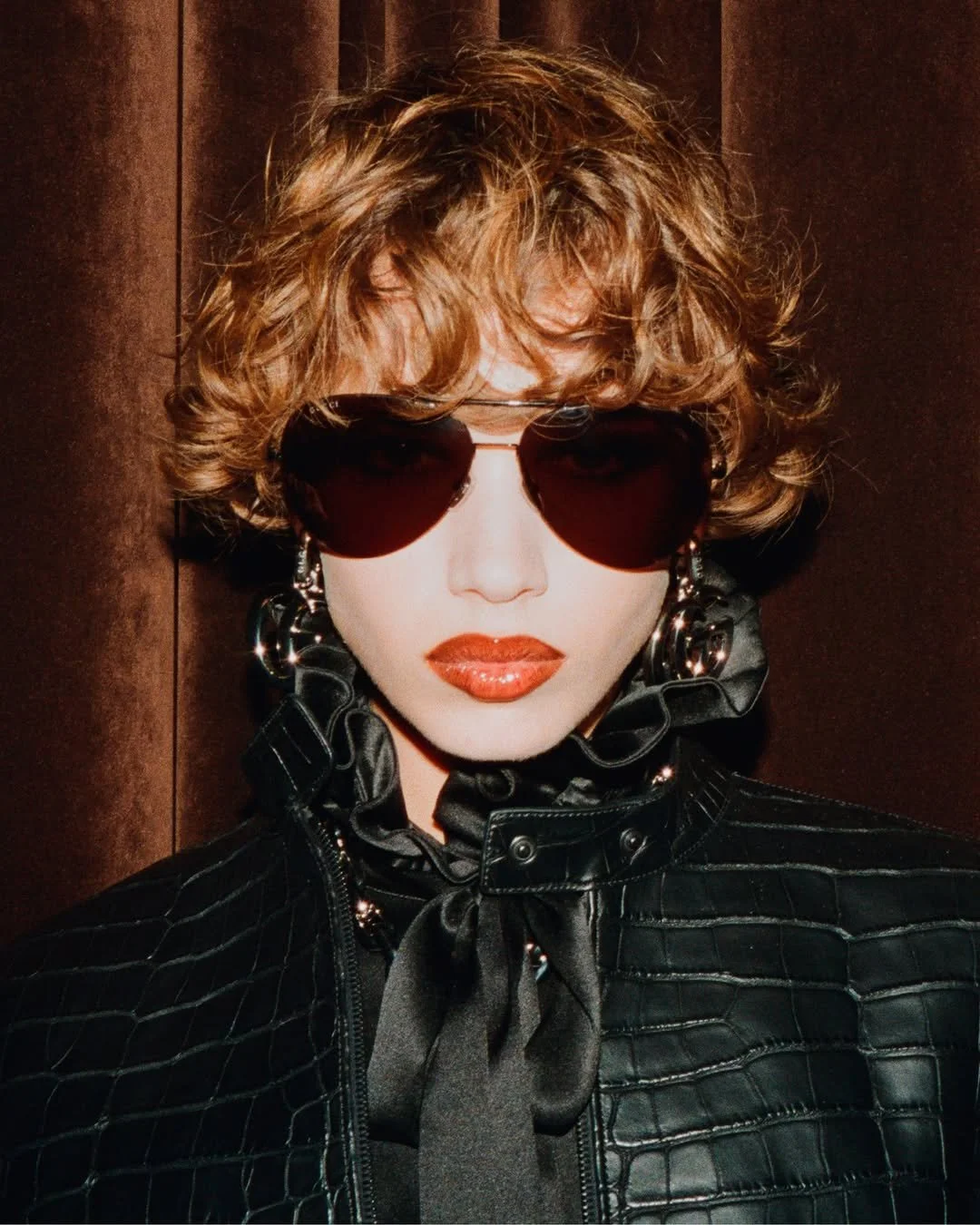 Une femme portant des lunettes de soleil foncées, un manteau noir en cuir avec un col volumineux, des boucles d'oreilles en forme de cercles et un ruban noir autour du cou, se tenant contre un mur en bois.