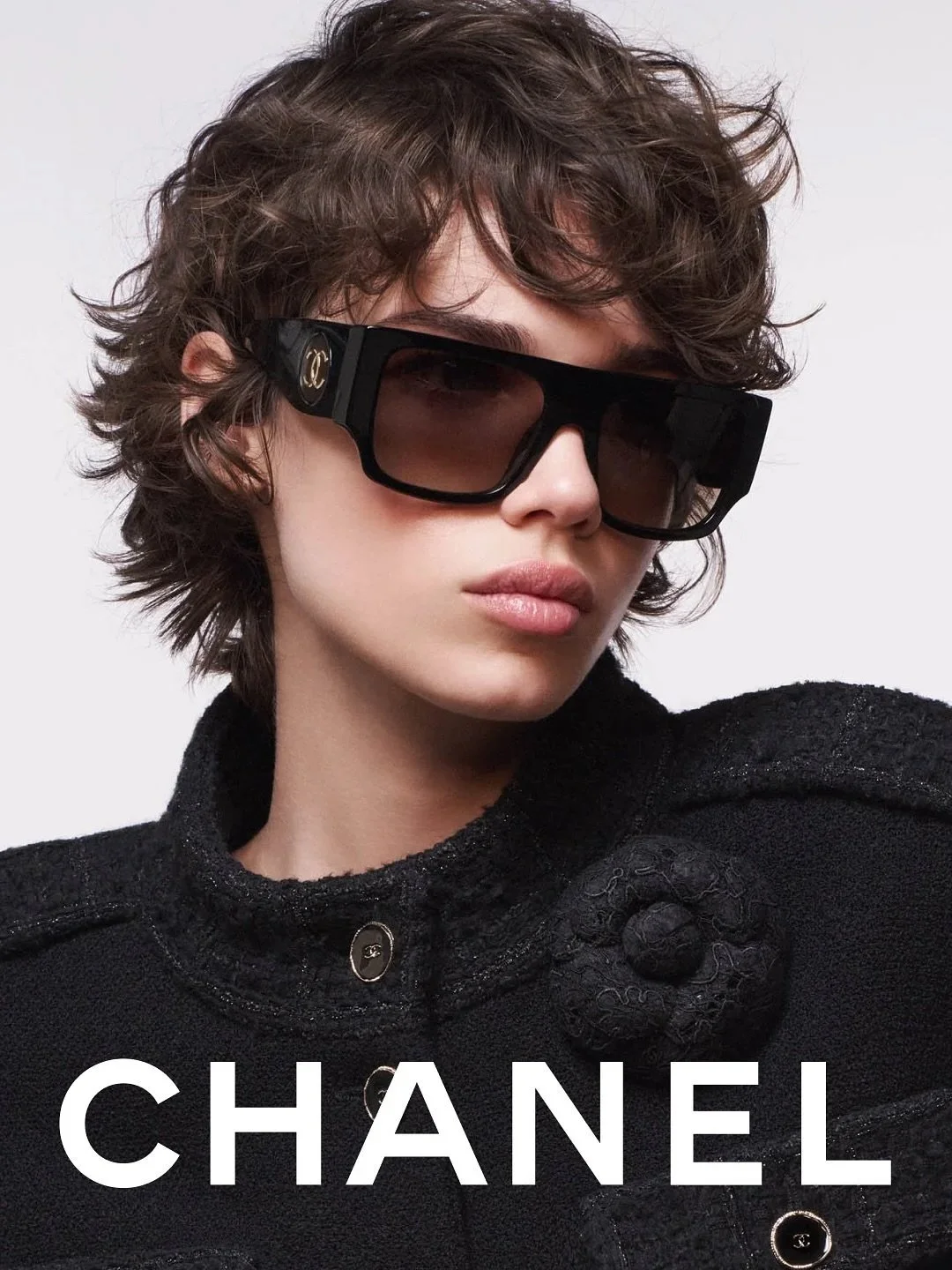 Une femme portant des lunettes de soleil noires de marque Chanel, avec un vêtement noir texturé, sur un fond blanc.