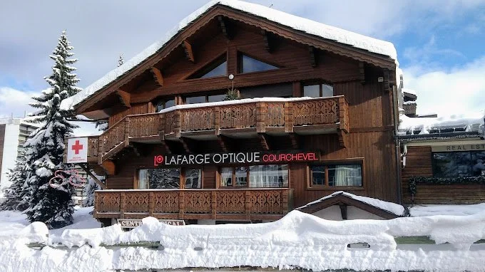 Bâtiment en bois avec un grand balcon, entouré de neige, avec une enseigne rouge 'LAFARGE OPTIQUE COURCHEVEL' et un signe de la Croix-Rouge.