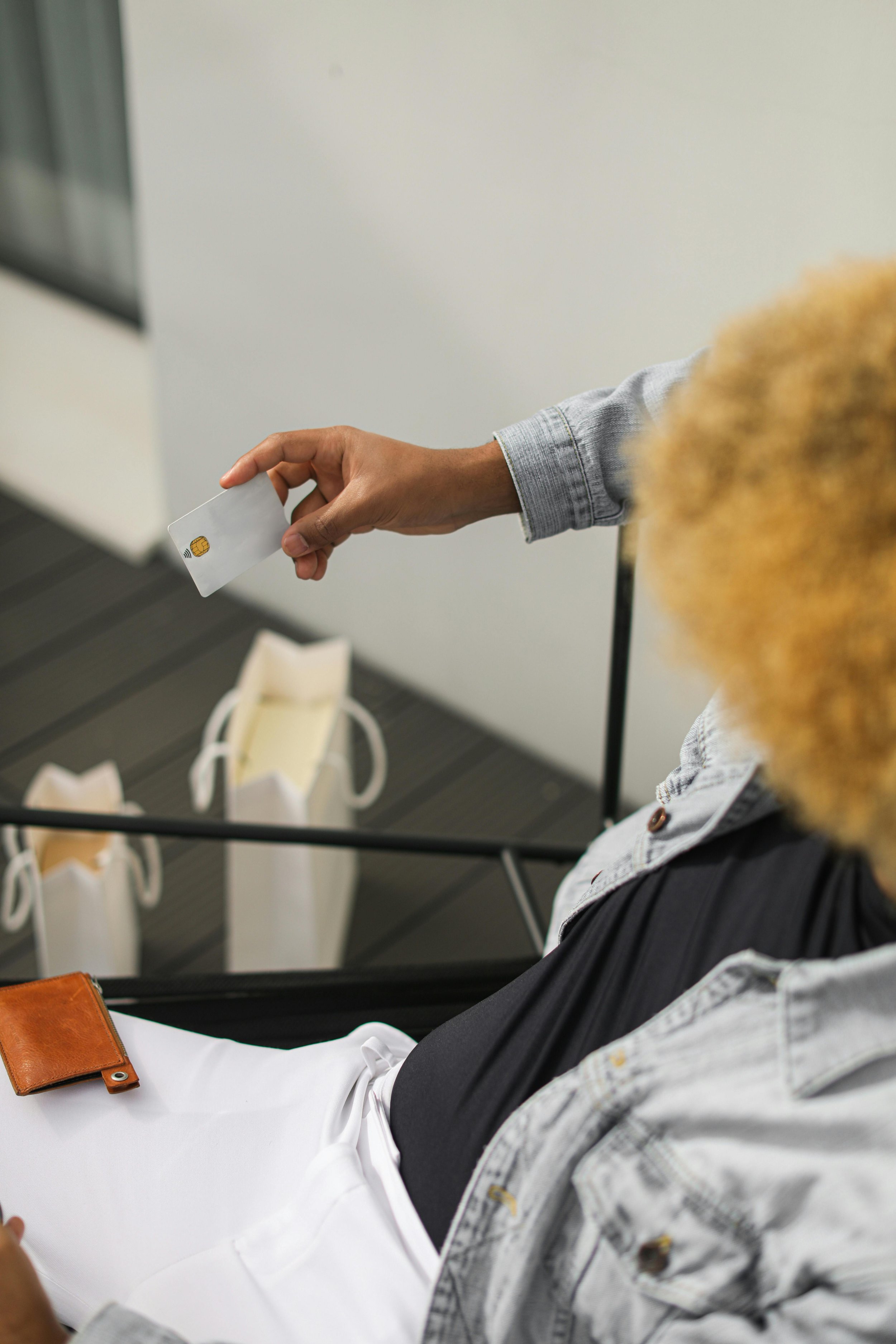 Une personne tenant une carte de crédit, assise avec une poche en cuir sur la jambe, avec d'autres sacs à main en arrière-plan.
