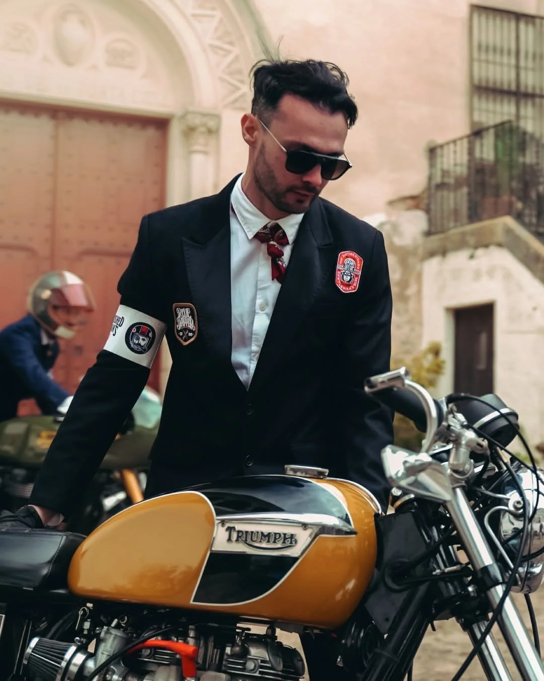 Un homme portant des lunettes de soleil et une veste noire avec des écussons, debout à côté d'une moto Triumph de couleur jaune-orange, dans un environnement extérieur avec un bâtiment en arrière-plan.