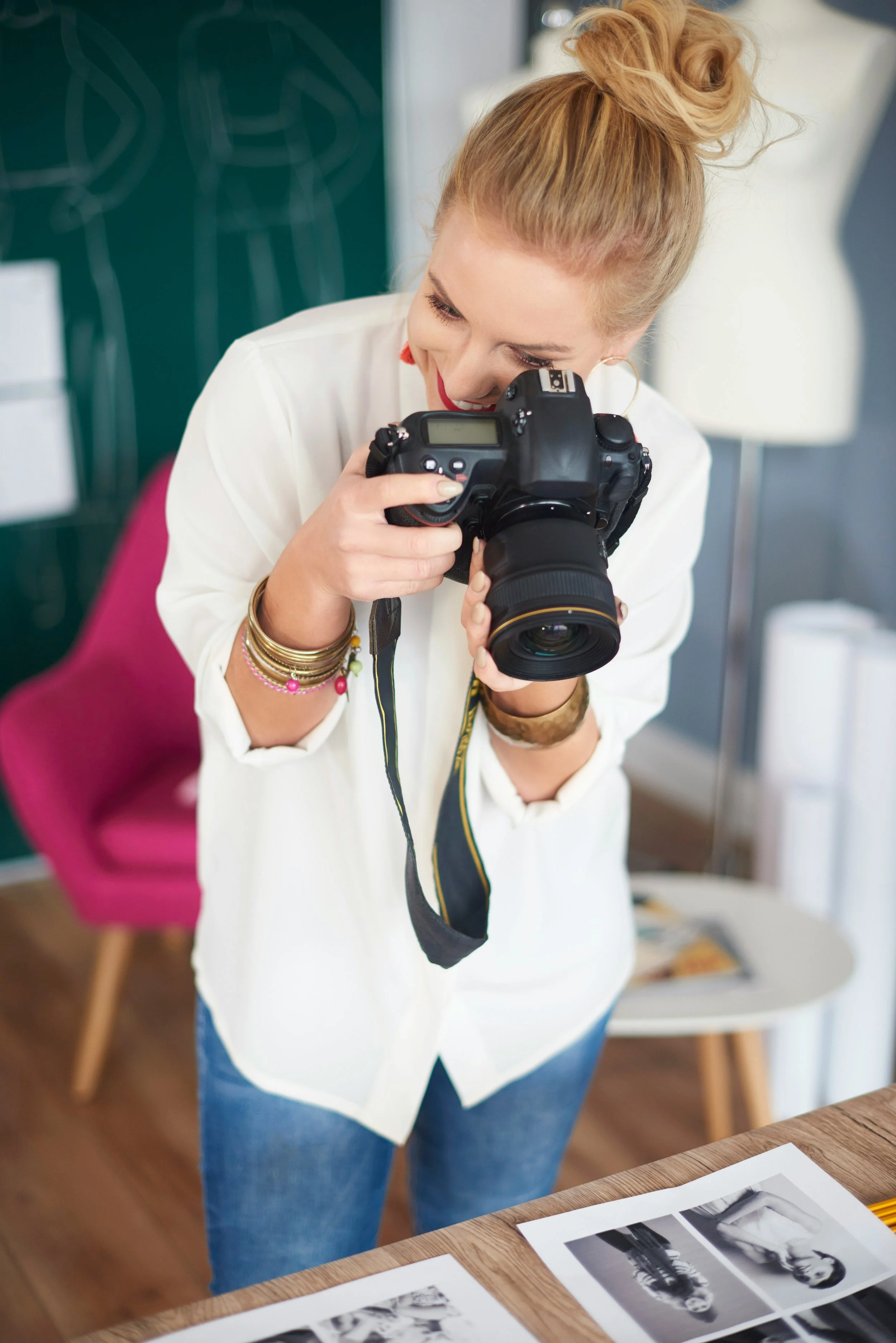 A woman with red hair tied in a bun is looking through a DSLR camera. She is wearing a white shirt, blue jeans, and jewelry on her hand and wrist. There are black-and-white photographs arranged on a wooden table in front of her, and a mannequin is visible in the background.