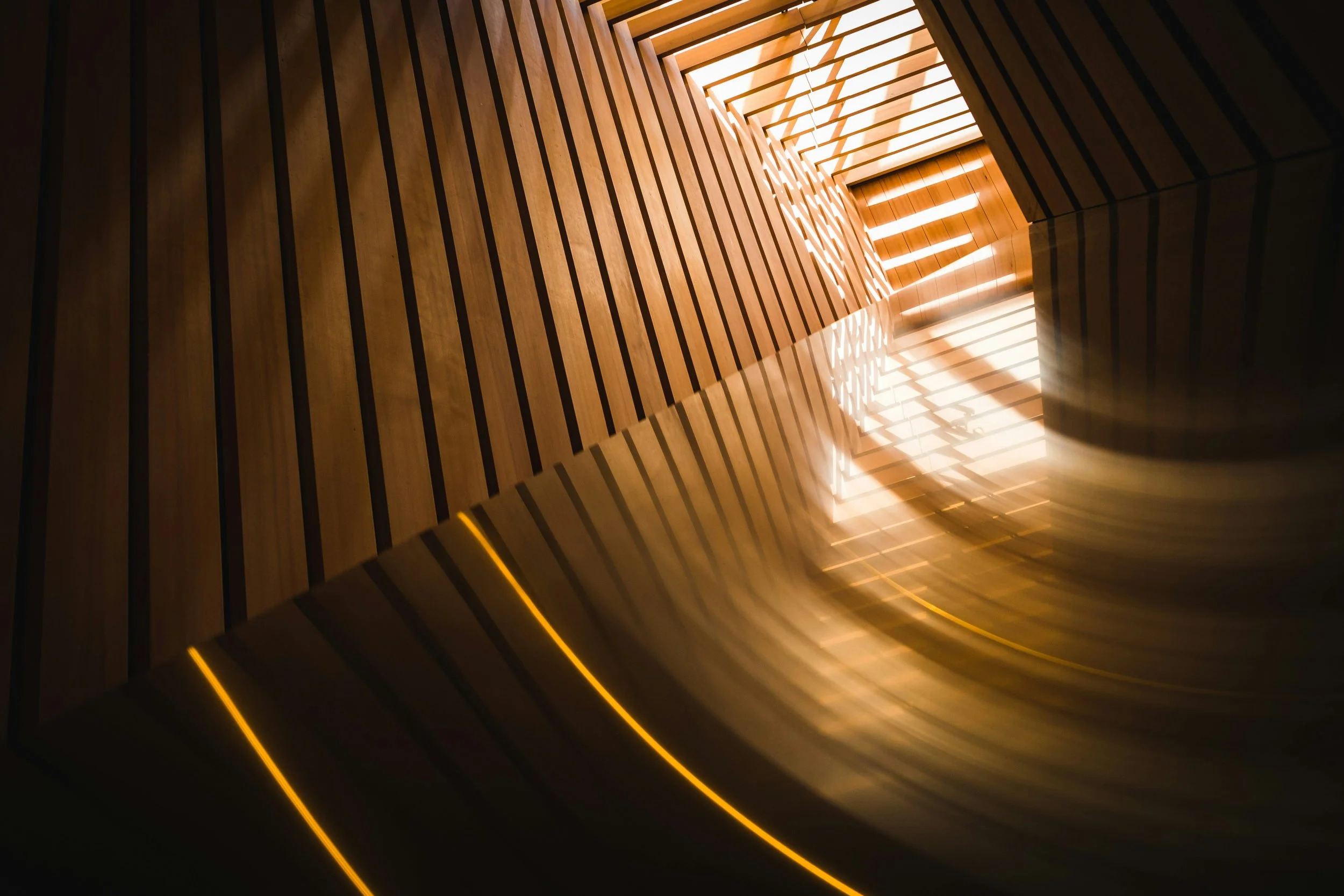 Interior view of a spiral staircase with wooden slats and warm lighting, looking upward toward a skylight.