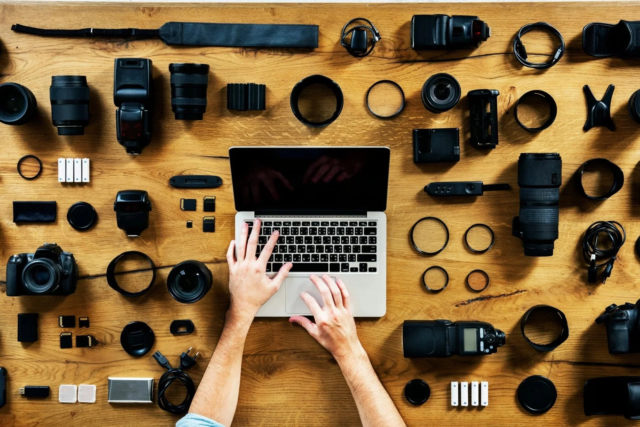A wooden table with various camera equipment, lenses, camera bodies, batteries, memory cards, and a laptop. Two hands are interacting with the laptop keyboard.