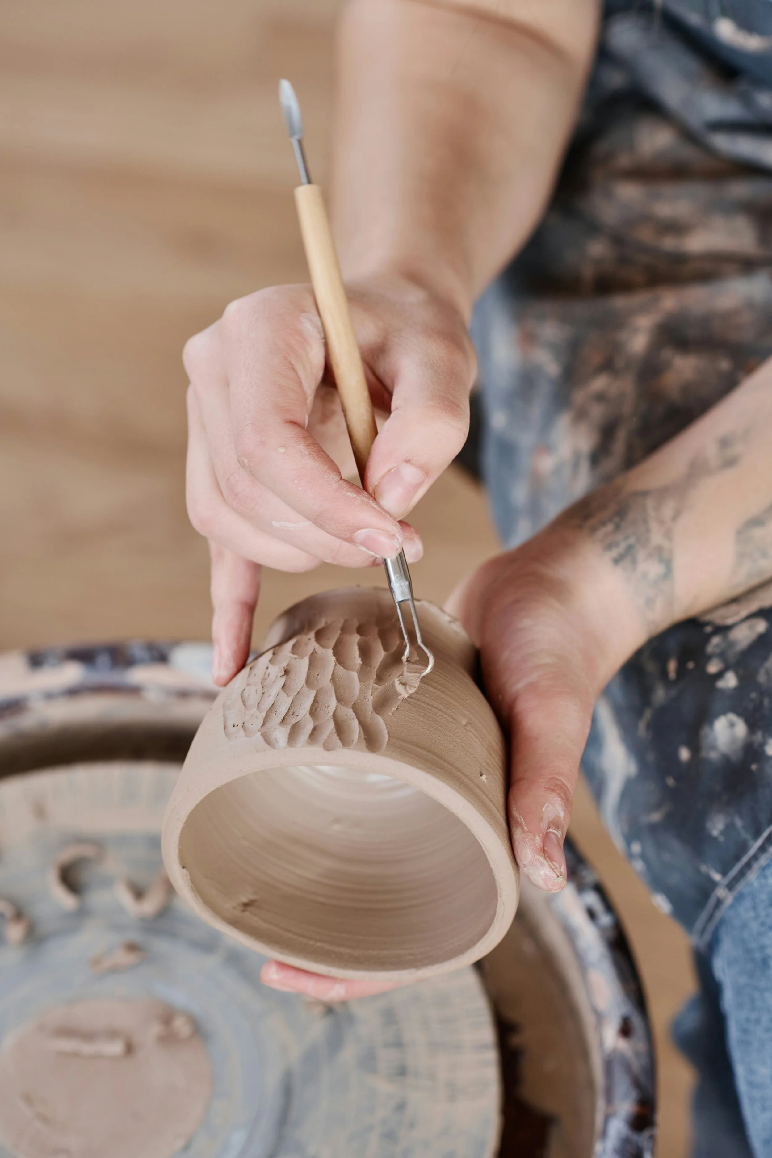 Close-up of person carving patterns into a ceramic bowl with a metal tool, wearing a shirt with camouflage pattern