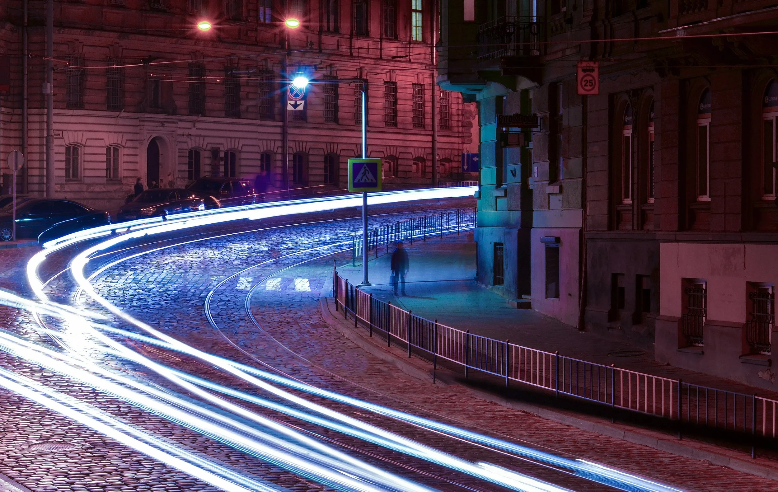 Nighttime city street scene with light trail streaks from moving vehicles, parked cars, old buildings, street signs, and streetlights.