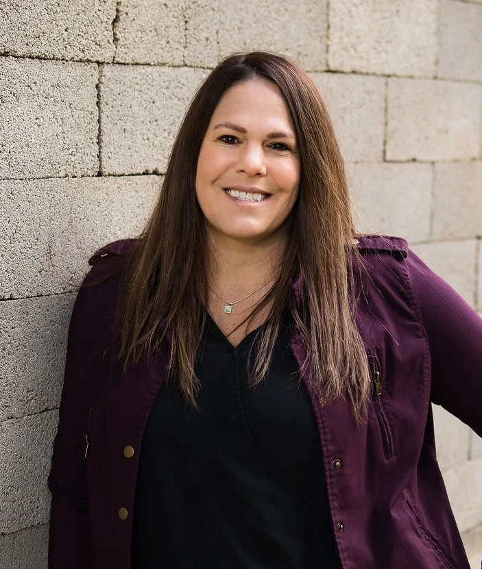 A woman with long brown hair smiling, wearing a black shirt and a purple jacket, standing against a beige brick wall.