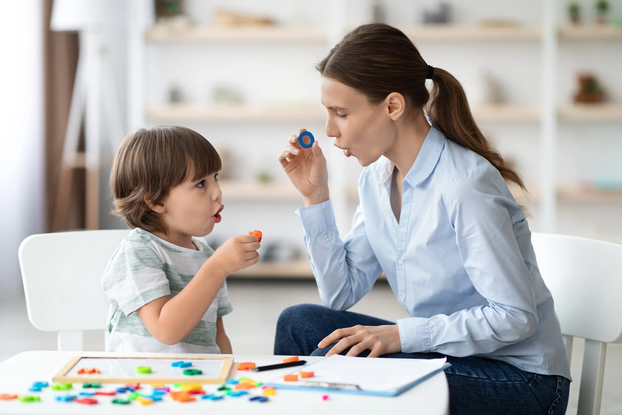 A woman and a young boy sit at a table playing with colorful letter tiles, with the woman blowing on a letter tile as the boy speaks and holds a letter piece.