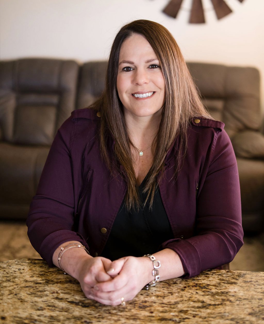 A woman with long brown hair, wearing a maroon jacket and jewelry, sitting at a granite table, smiling at the camera in a living room with a brown couch and a wall decoration.