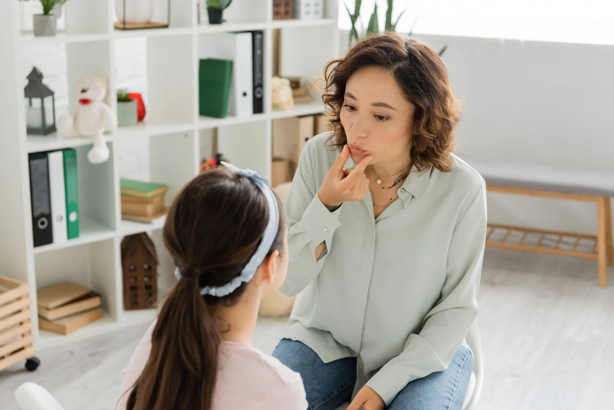 A woman with curly brown hair and wearing a light-colored blouse is sitting across from a young girl with long brown hair and a headband. They are in a room with a white bookshelf and a gray cushioned bench in the background, engaging in a serious conversation.