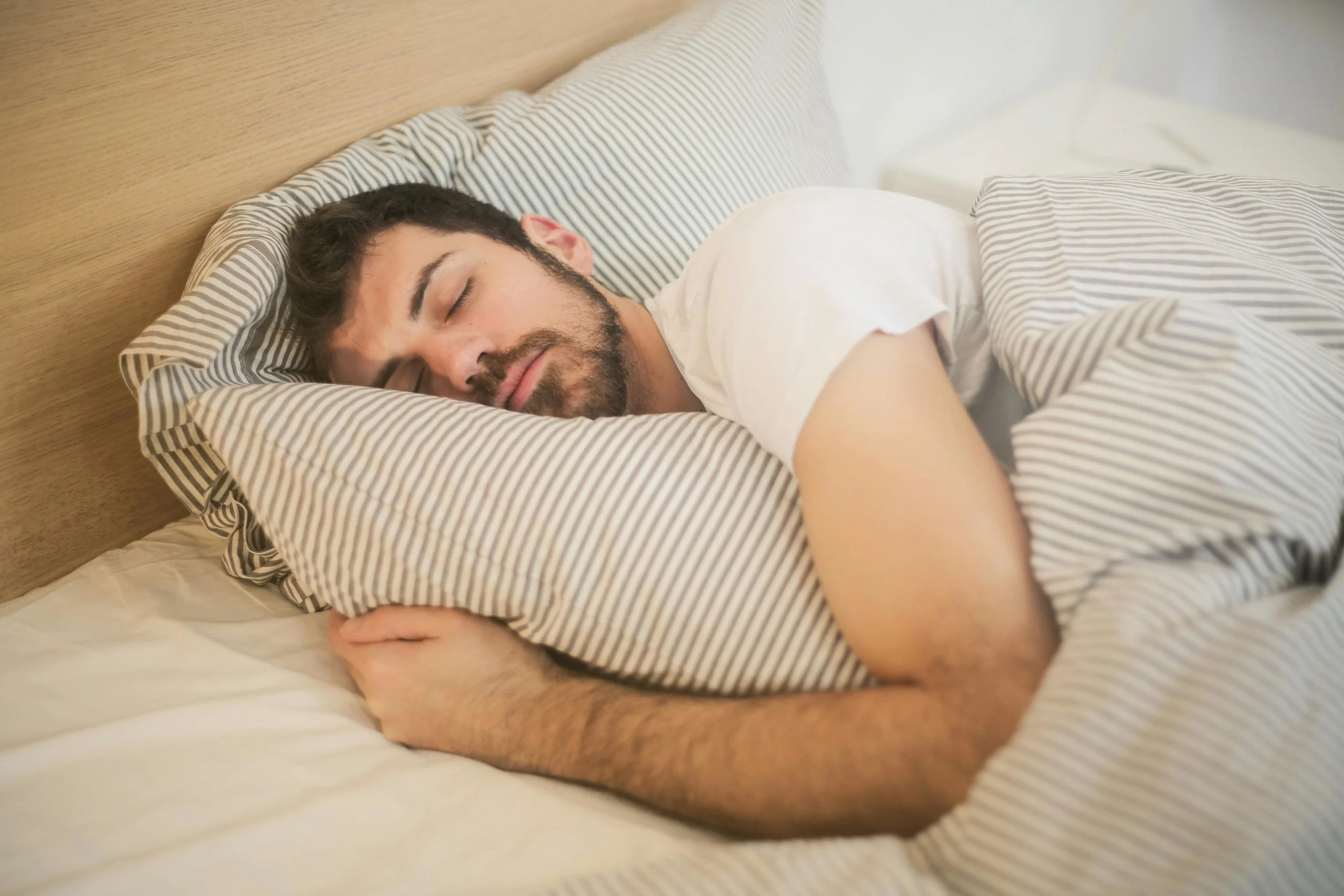 A man with dark hair and a beard sleeping peacefully on a bed with striped pillowcases and a matching blanket.