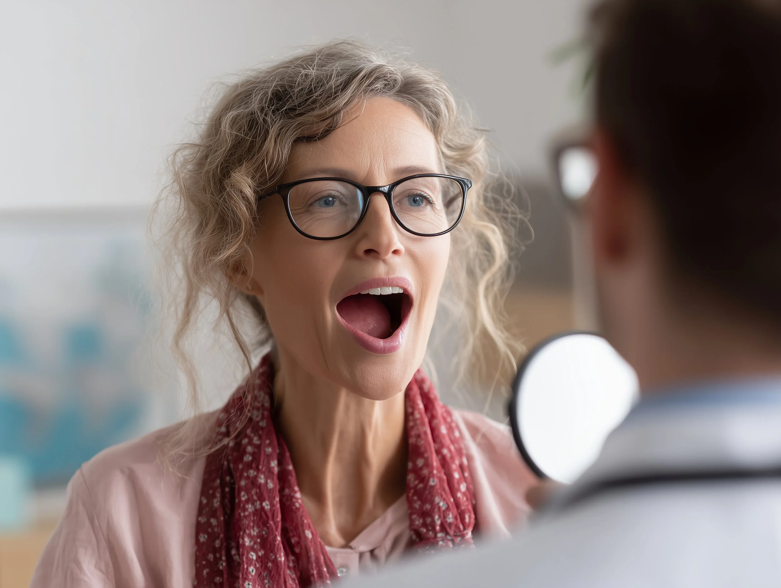 A middle-aged woman with curly blonde hair, glasses, and a pink shirt, appears surprised or shocked while talking to a doctor in a medical office.