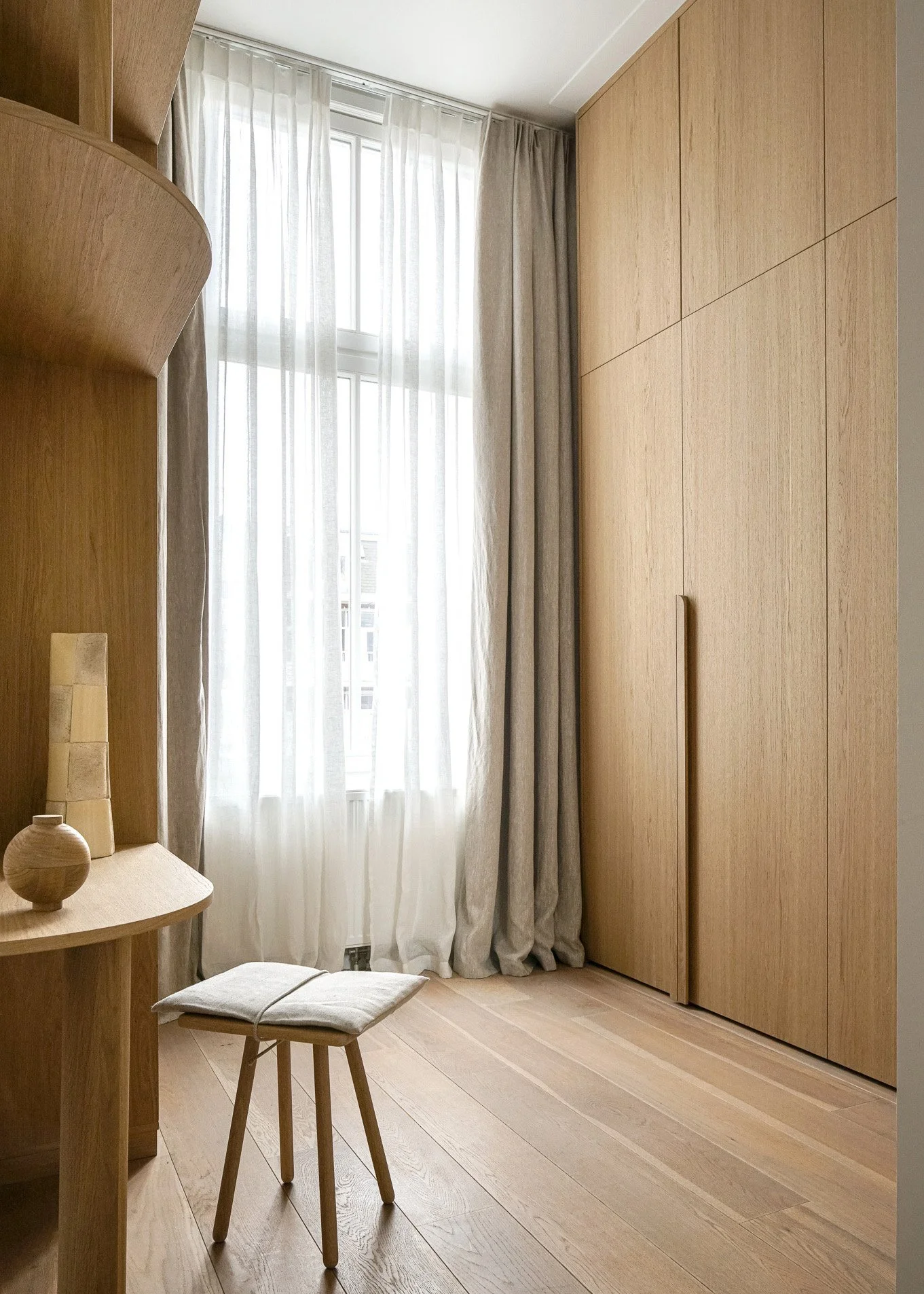 A minimalist room with wooden furniture and flooring, a window with sheer curtains, and a small stool near a desk with decorative objects.