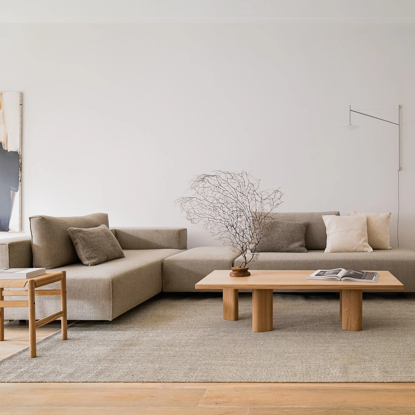 Minimalist living room with beige sectional sofa, neutral pillows, wooden coffee table with a decorative dried leaf branch, and light-colored walls with artwork and wall-mounted lamp.