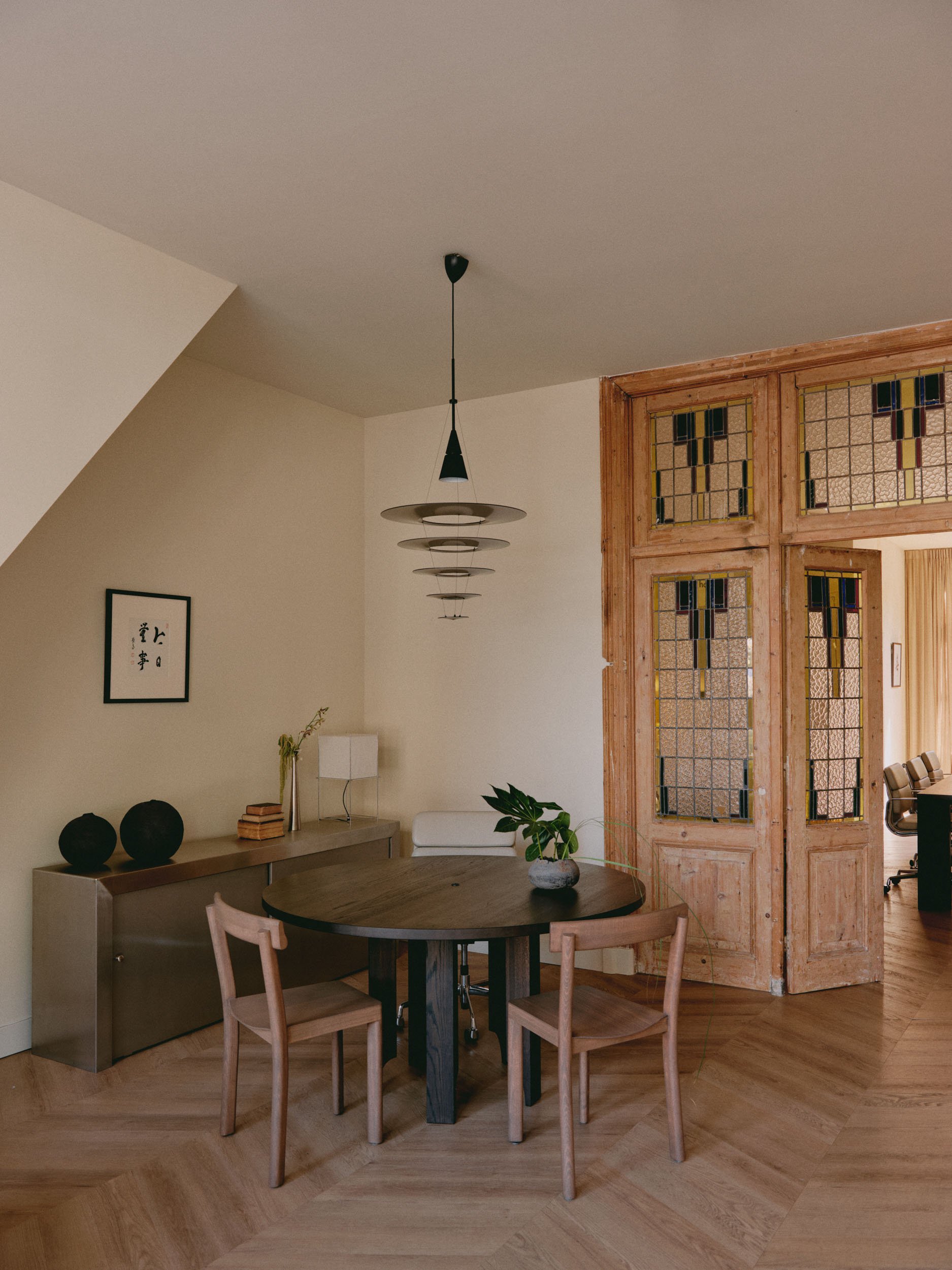 A cozy dining area with a round wooden table, three matching wooden chairs, and a decorative centerpiece. The room features a modern hanging light fixture, a sideboard with black vases and a framed artwork on the wall, and a partially open wooden lattice door leading to an adjoining space with a desk and chairs.