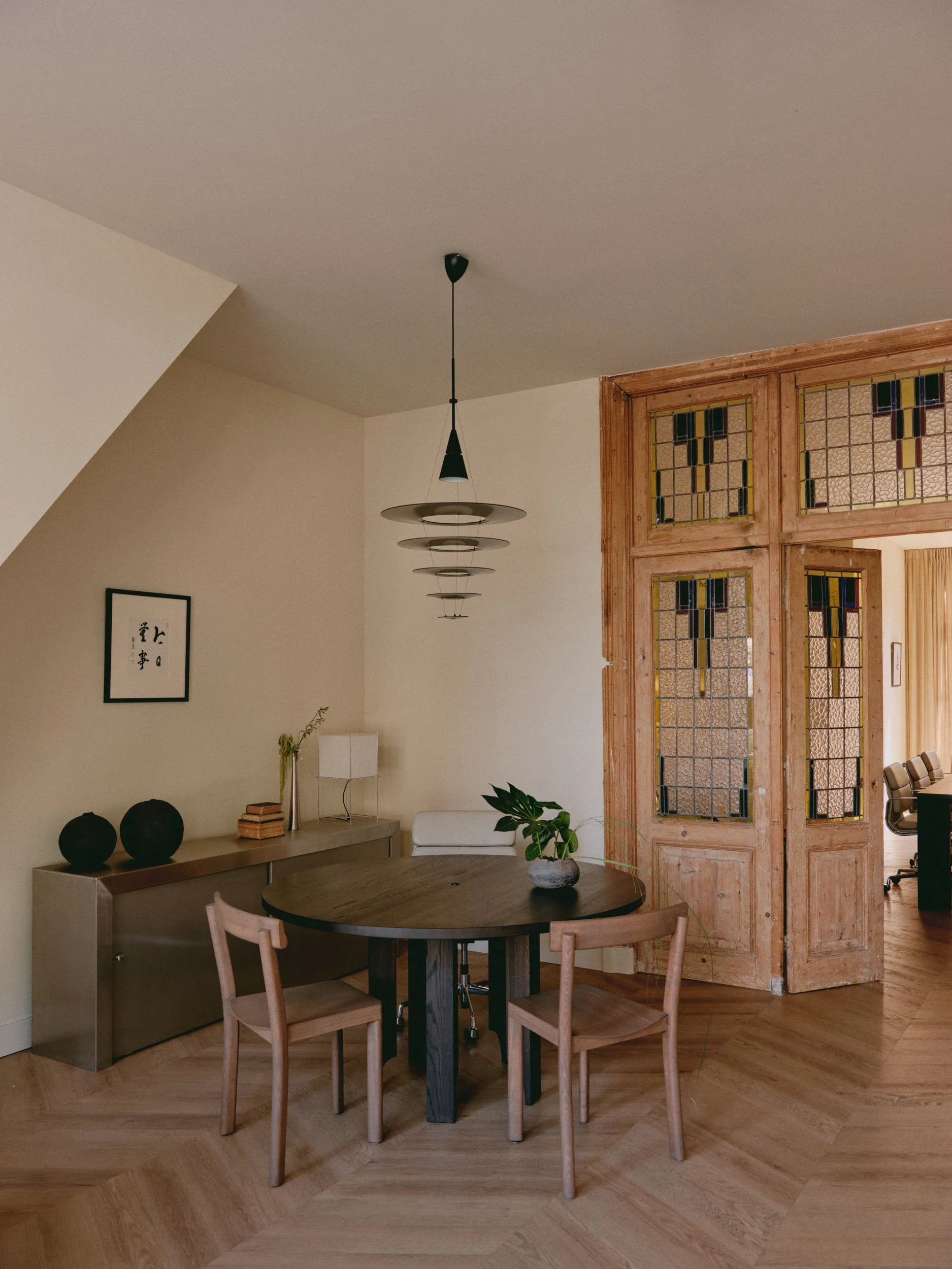 Interior of a room featuring a round wooden dining table with three chairs, a sideboard with decorative objects and a lamp, a modern hanging light fixture, a small plant, and wooden framed stained glass doors in the background.