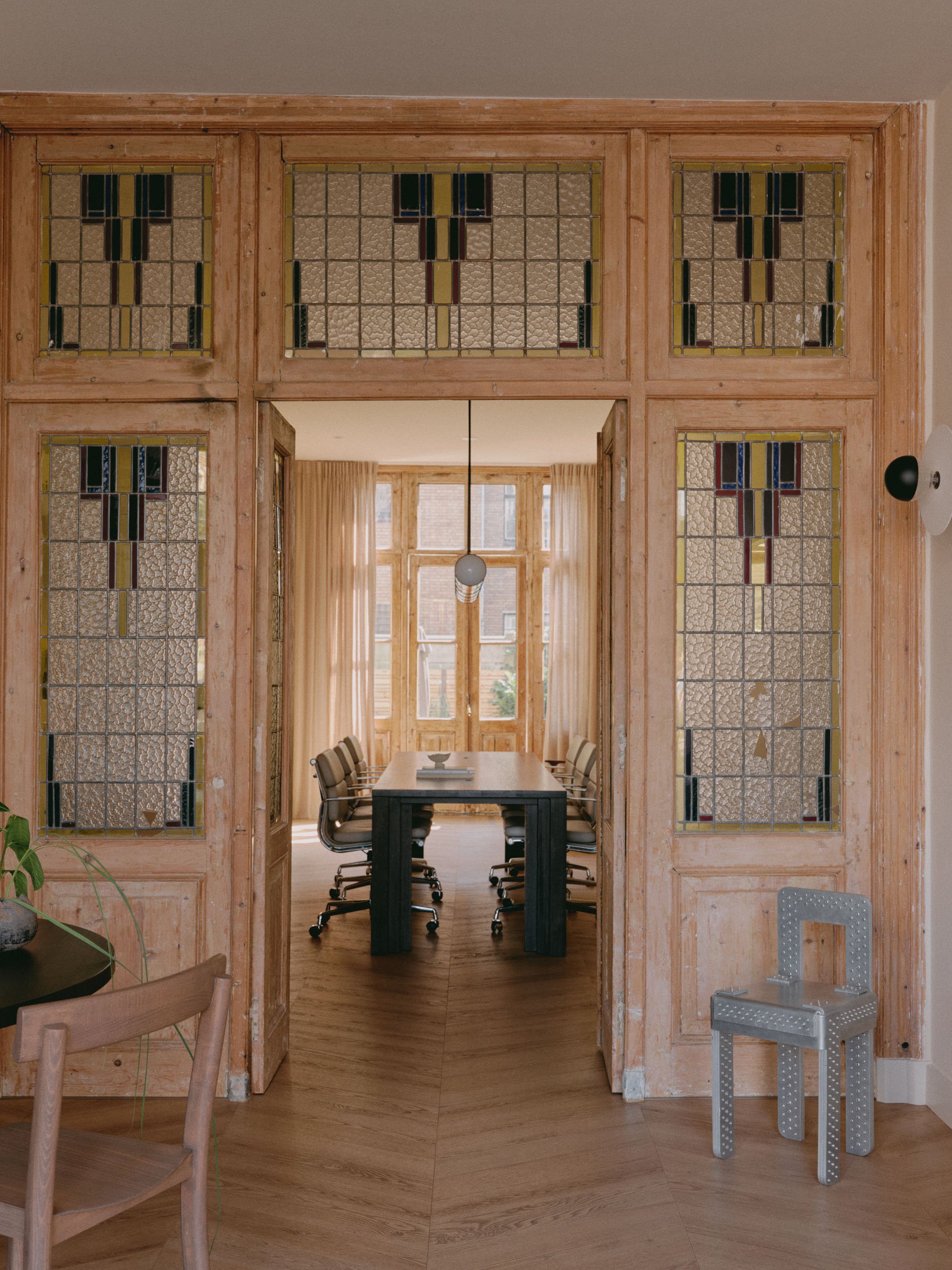 View of a dining room through a wooden-framed glass partition with stained glass accents. The room features a long table with chairs, large windows with curtains, and a hanging pendant light.
