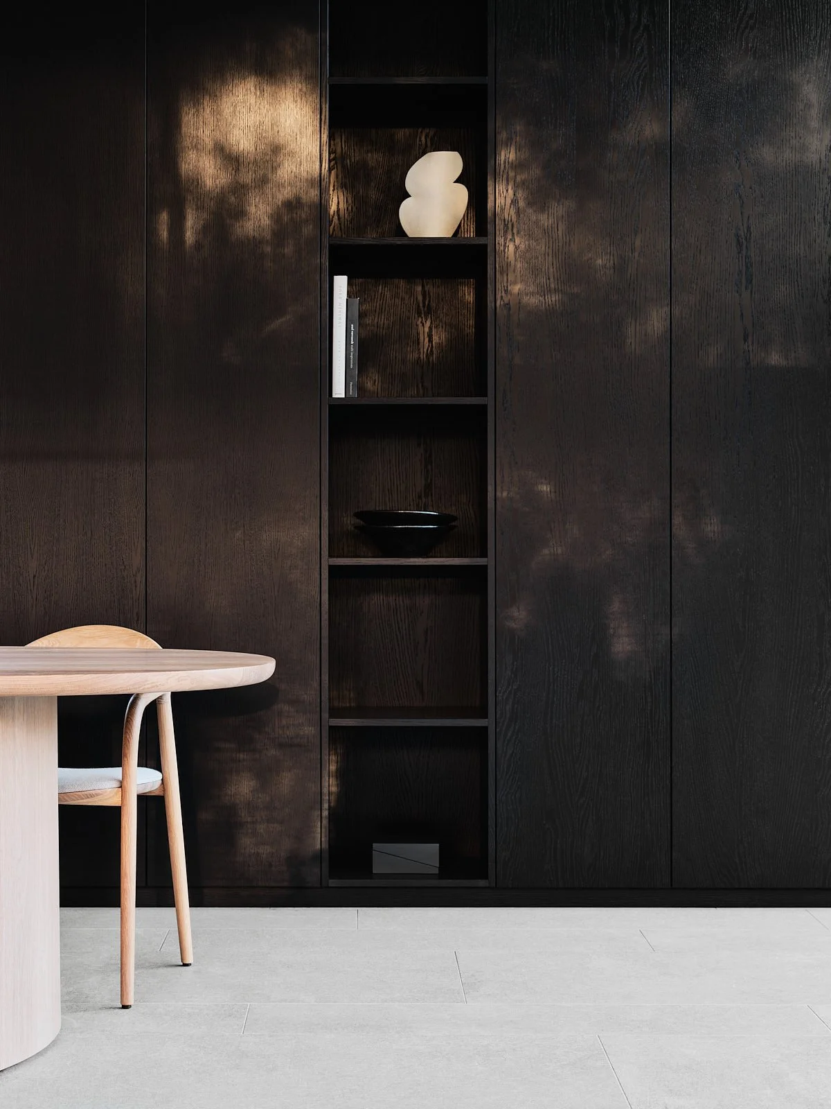 Interior scene with dark wooden wall unit and a small, round, light-colored wooden table with matching chair, against a smooth, light-colored tiled floor.