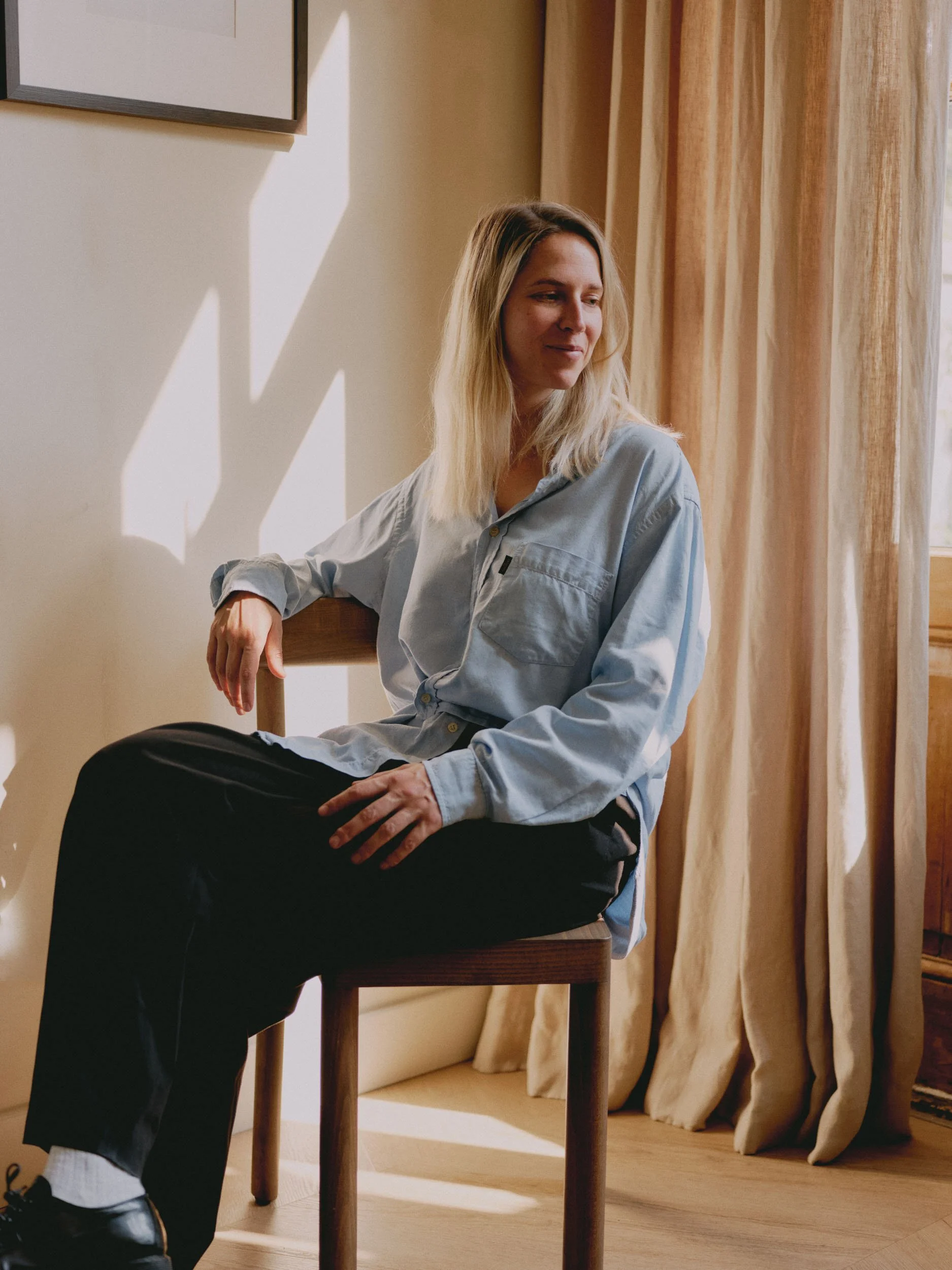 A young woman with blonde hair sitting on a wooden chair near a window with beige curtains, looking thoughtfully to the side.