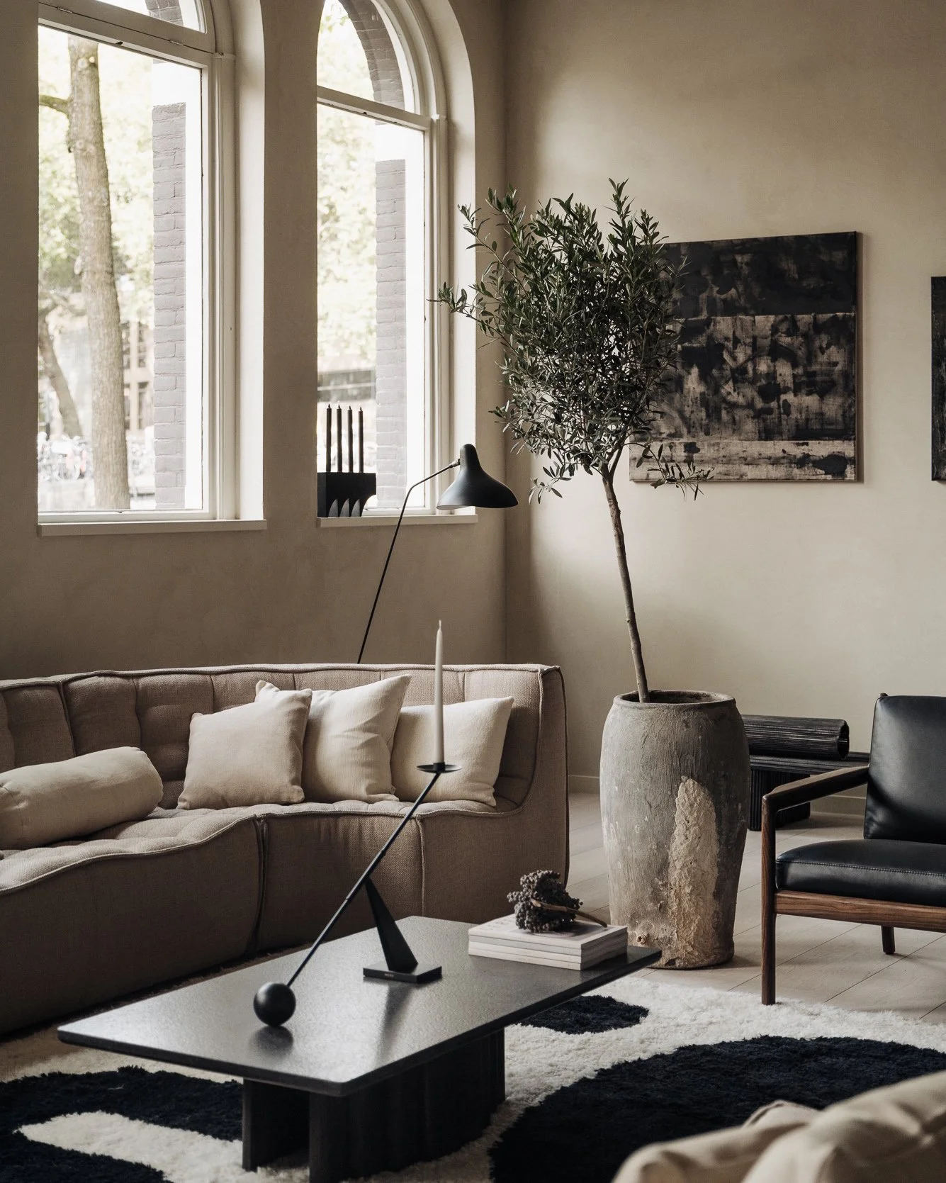 Modern living room with a beige tufted sofa, a black armchair, large window with arched top, potted plant next to the window, abstract art on the wall, black coffee table with decorative objects, floor lamp, and a black and white rug.