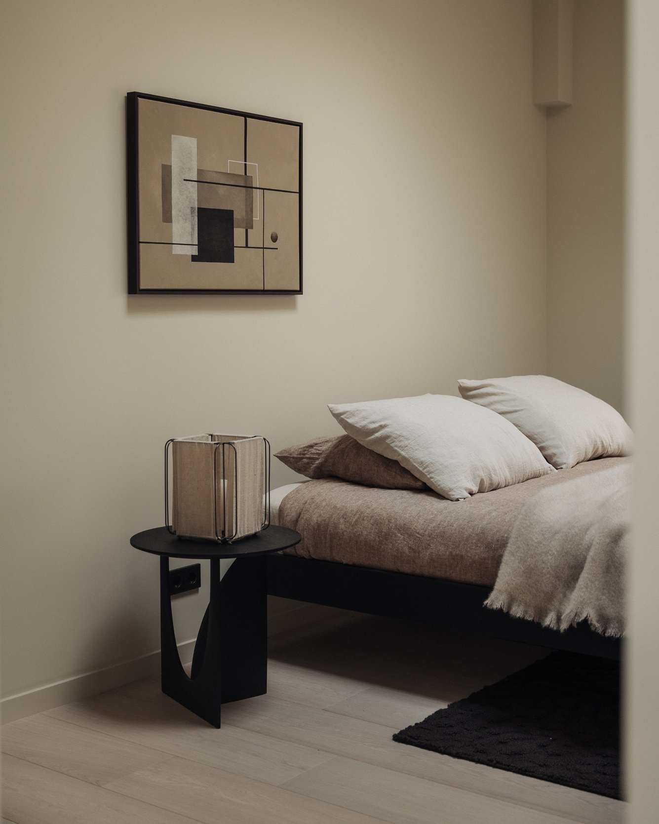 Bedroom with a bed, beige bedding, and three white pillows, black side table with a modern lamp, abstract wall art, and a black rug on light-colored wooden floor.