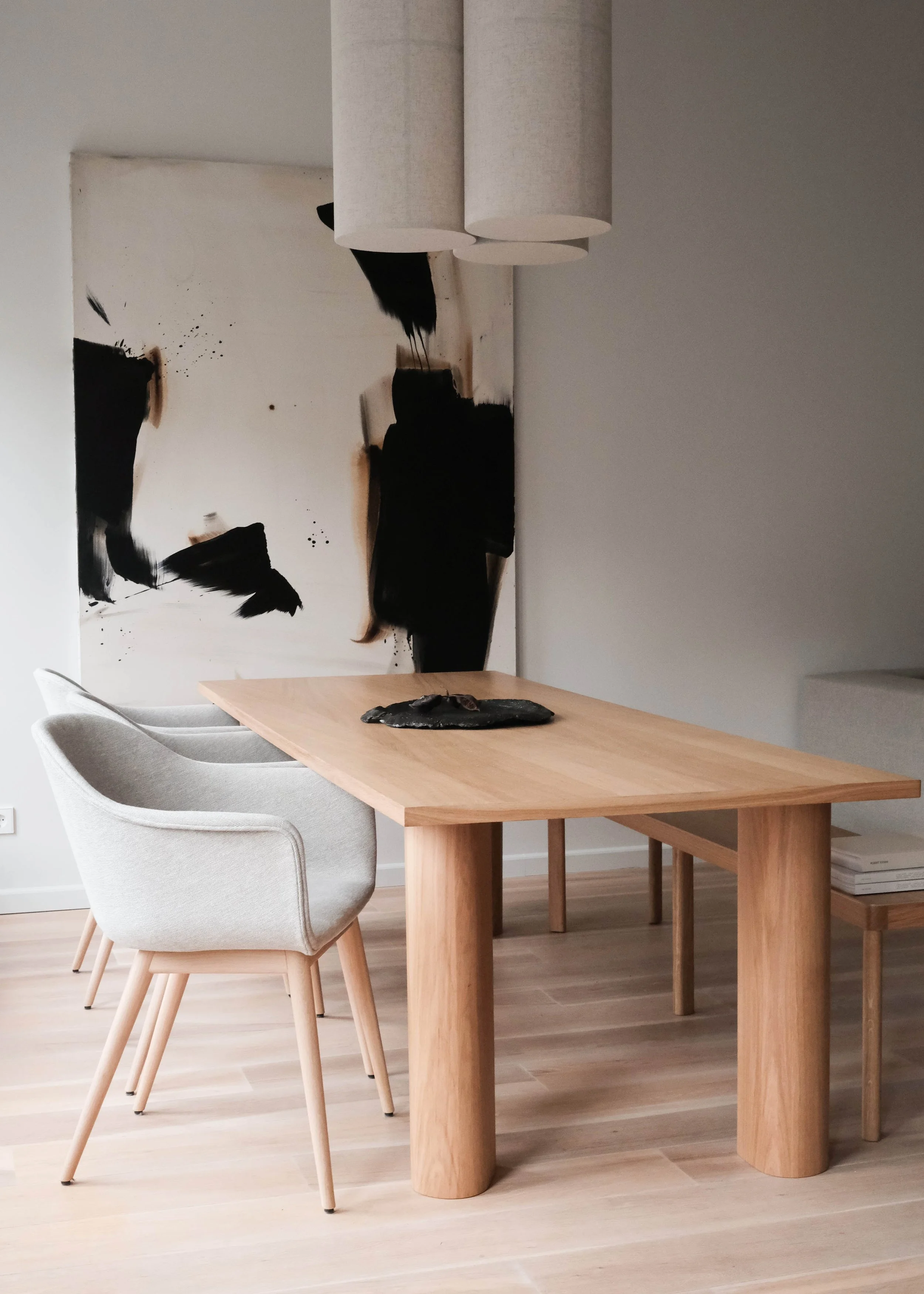 A minimalist dining room with a light wooden table, two white upholstered chairs, abstract black and white artwork on the wall, and a black sculpture or decoration on the table.